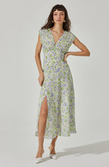 A woman is wearing a long, flowy dress with a floral pattern in shades of purple, green, and white. The dress has a V-neckline and short, slightly caped sleeves. It features a wrapped bodice and a high slit on the left side, revealing her leg. The fabric appears lightweight and semi-transparent. She is also wearing white shoes with a bow detail.