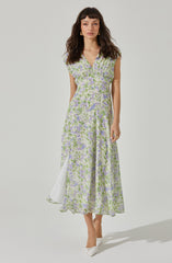 A woman is wearing a sleeveless, ankle-length dress with a V-neckline. The dress features a light purple and green floral pattern on a sheer fabric, with a white lining underneath. The dress has a front slit that reveals a white inner layer. She is also wearing white pointed-toe shoes with a bow detail.