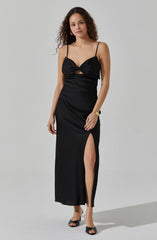 A woman is wearing a black spaghetti strap dress with a ruched bust that features a bow detail and a keyhole cutout below the bust. The dress is fitted at the waist and has a high slit on one side, revealing part of her leg. She pairs the dress with black open-toe heels and gold bangles on her wrist.