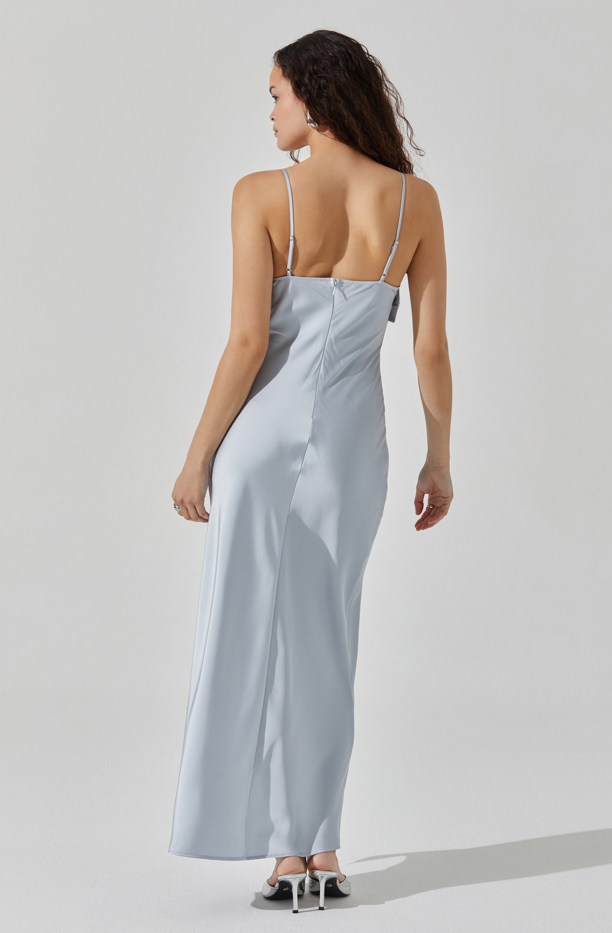 A woman is wearing a long, satin, light blue dress with thin spaghetti straps. The dress has a fitted bodice and flows down to the floor, creating an elegant silhouette. She is also wearing high-heeled shoes that are black and white.