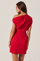 A woman is wearing a fitted, short red dress with an asymmetrical neckline that drapes over one shoulder, creating a loose sleeve. The fabric appears to be a smooth, slightly shiny material, and the dress has a simple, elegant silhouette that accentuates her figure.