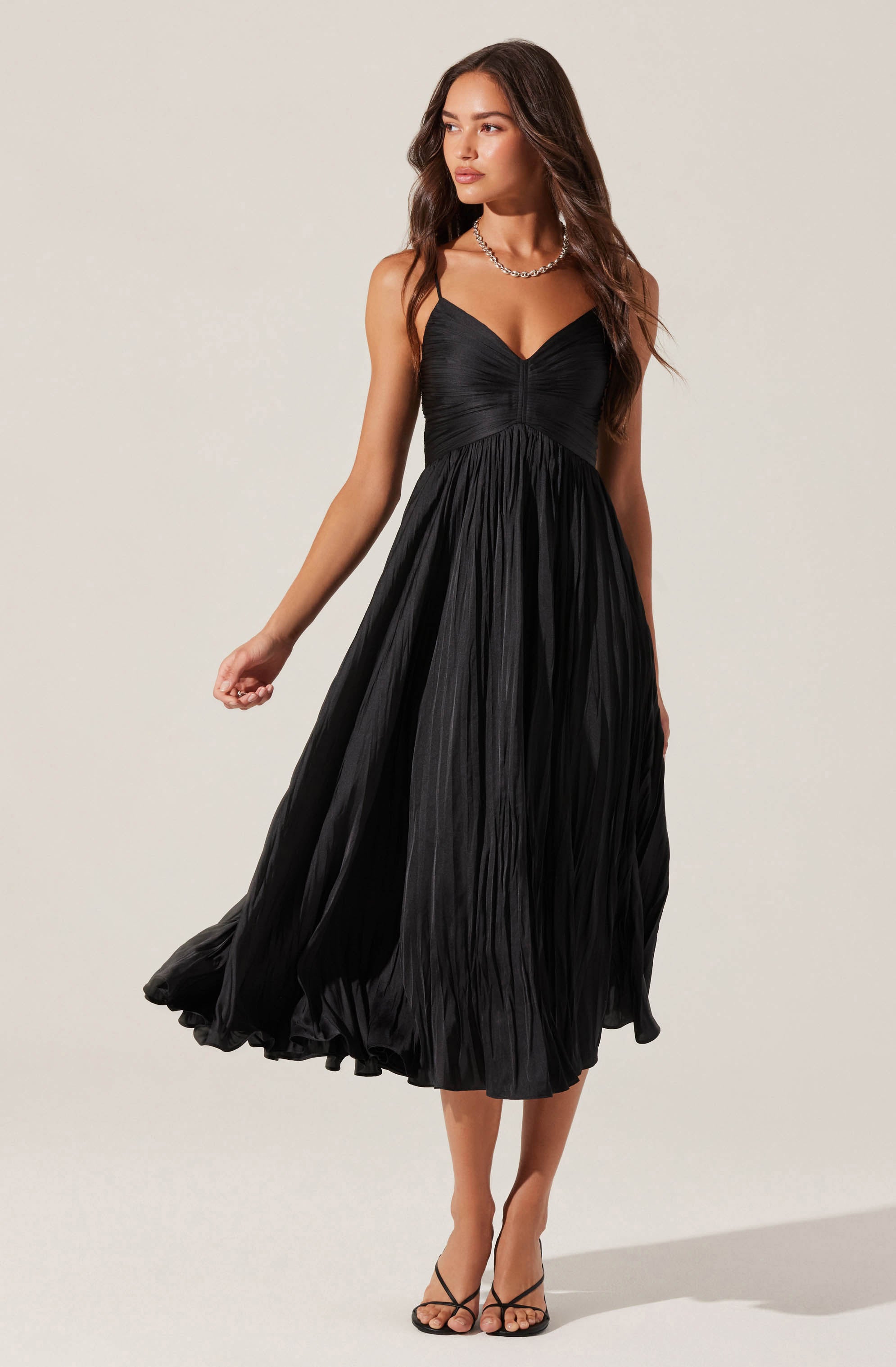 A woman is wearing a black pleated midi dress with thin spaghetti straps and a fitted bodice, paired with a silver necklace and black high-heeled sandals.