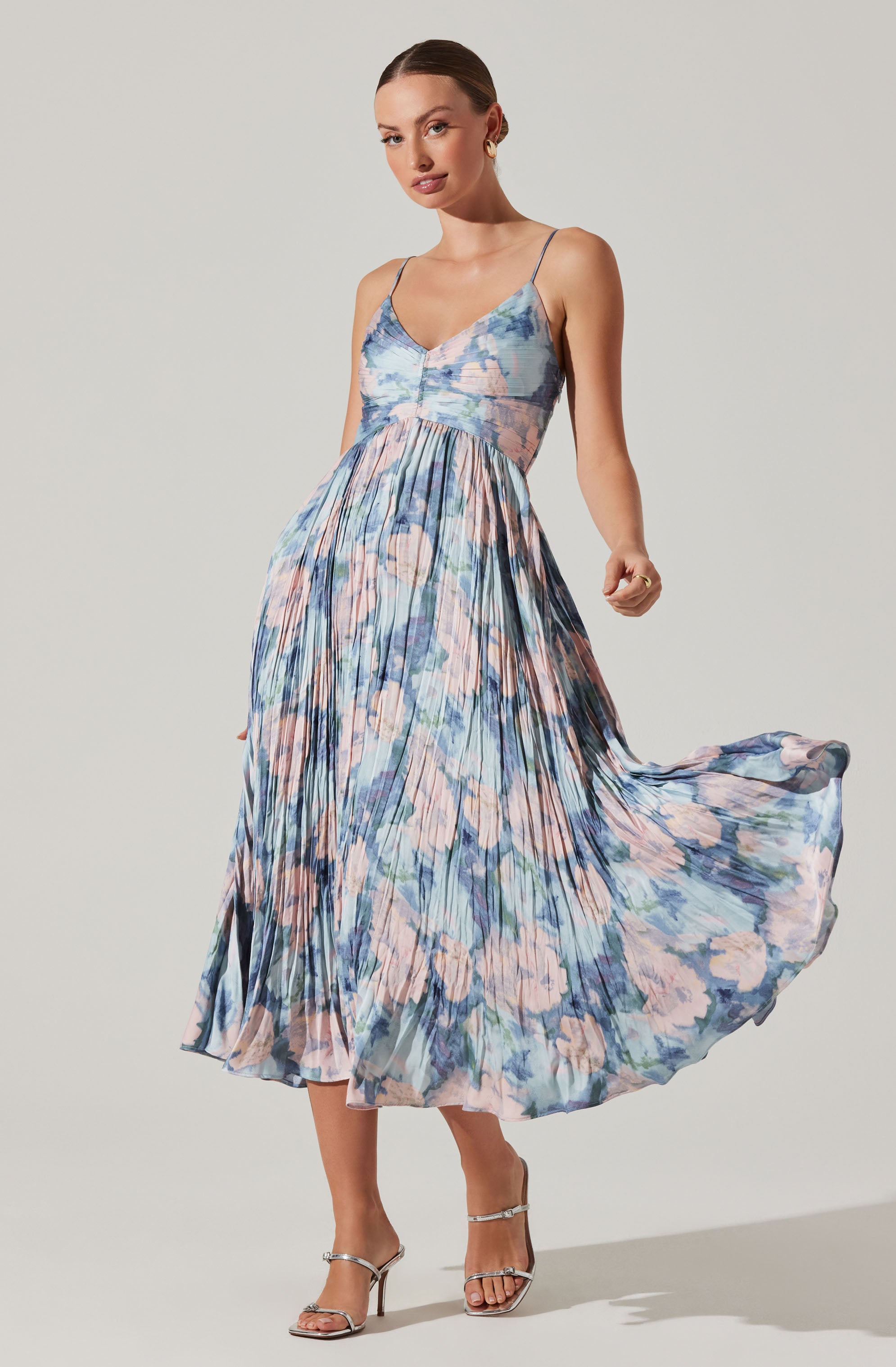 A woman is wearing a sleeveless, ankle-length dress with thin spaghetti straps. The dress features a fitted bodice and a full, pleated skirt with a floral pattern in shades of pink, blue, peach, and green. The fabric appears lightweight and flowy. She is also wearing silver high-heeled sandals with ankle straps.