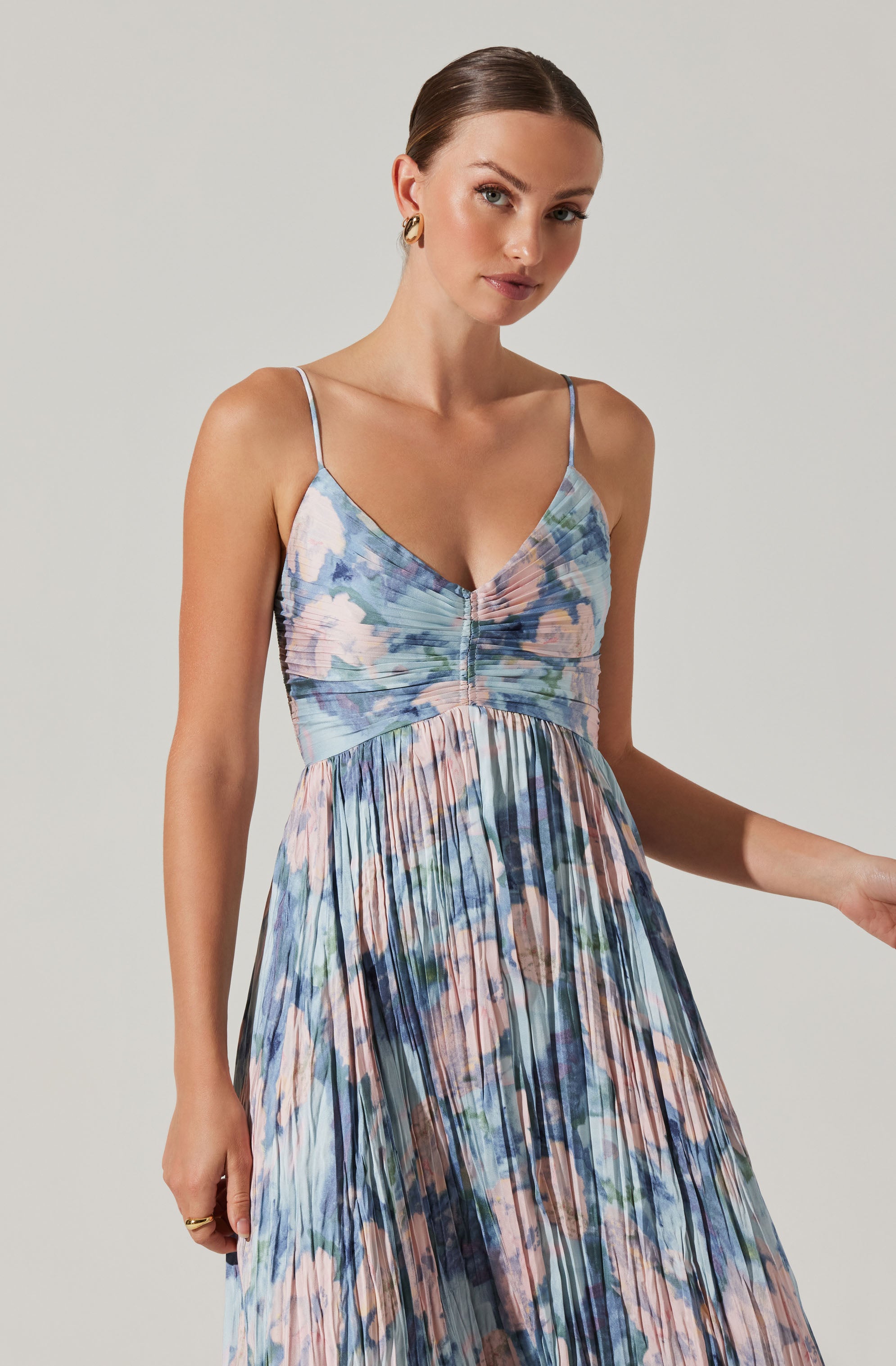 A woman is wearing a sleeveless dress with thin spaghetti straps. The dress features a fitted bodice with a shimmery, cinched detail at the center, and a flowing, pleated skirt styled with an abstract, watercolor-like pattern in shades of blue, pink, green, and cream. She has her hair pulled back and is accessorized with small hoop earrings.