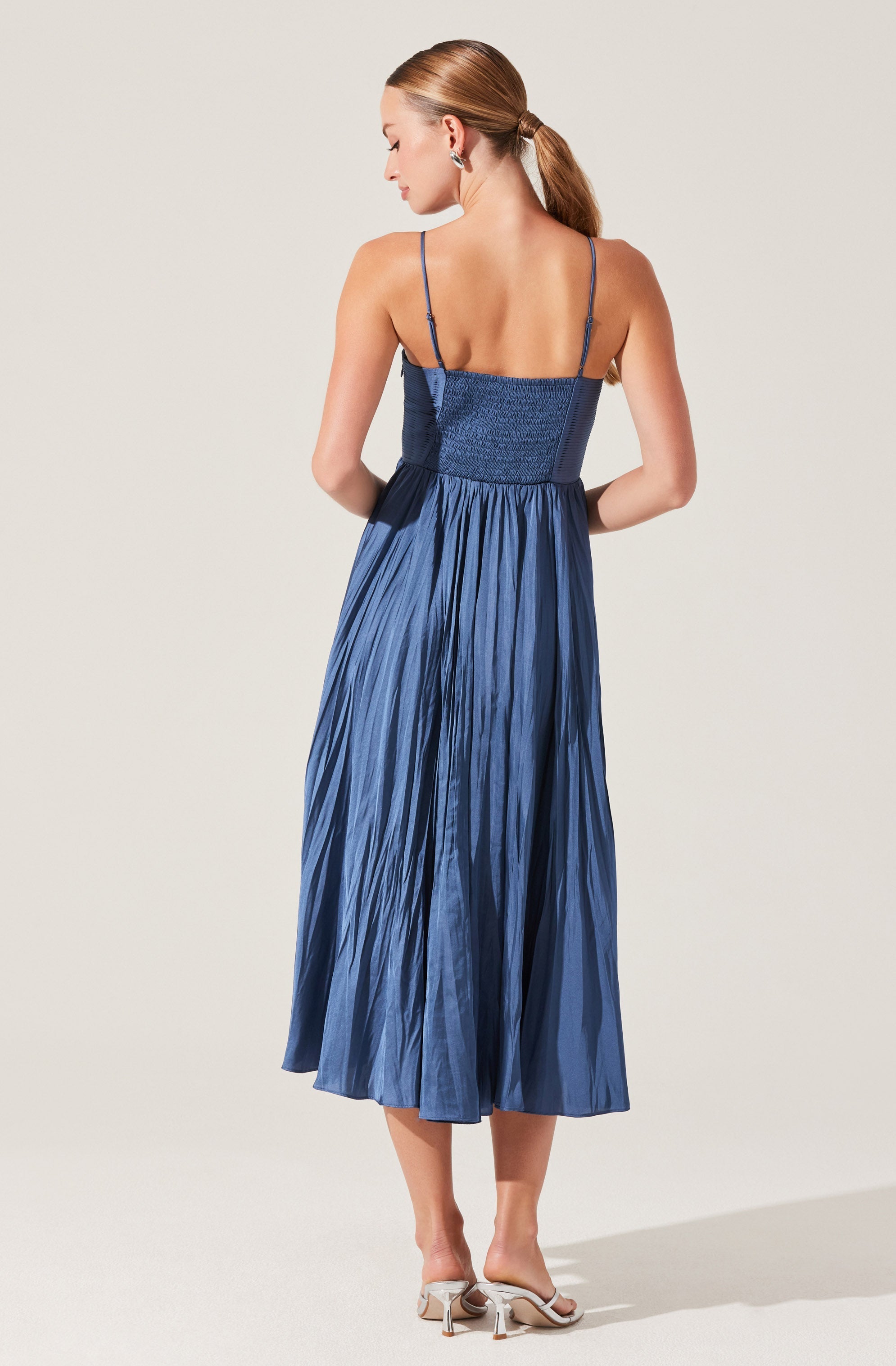 A woman is wearing a blue dress with thin spaghetti straps. The bodice is fitted and textured, while the skirt is long, pleated, and flowing. She is also wearing white high-heeled sandals.