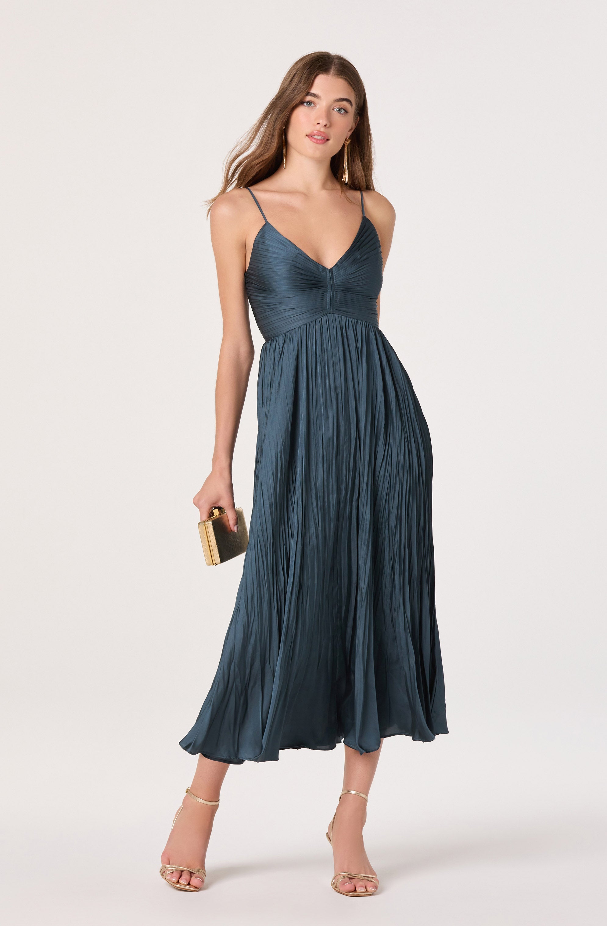 A woman is wearing a dark blue, pleated, sleeveless dress with thin spaghetti straps. The dress has a fitted bodice with a V-neckline and flows down to a midi length. She is holding a small, rectangular clutch and wearing strappy high-heeled sandals.