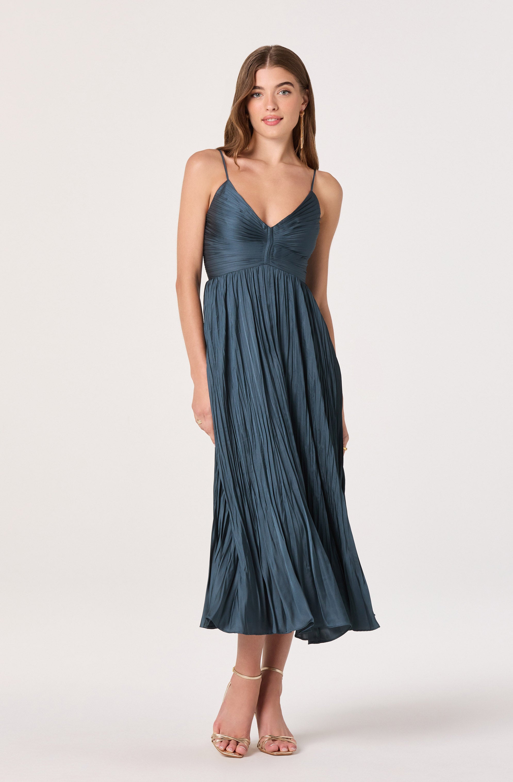 A woman is wearing a dark blue, pleated sundress with a fitted, ruched bodice featuring a V-neckline and thin spaghetti straps. She is also wearing strappy high-heeled sandals.