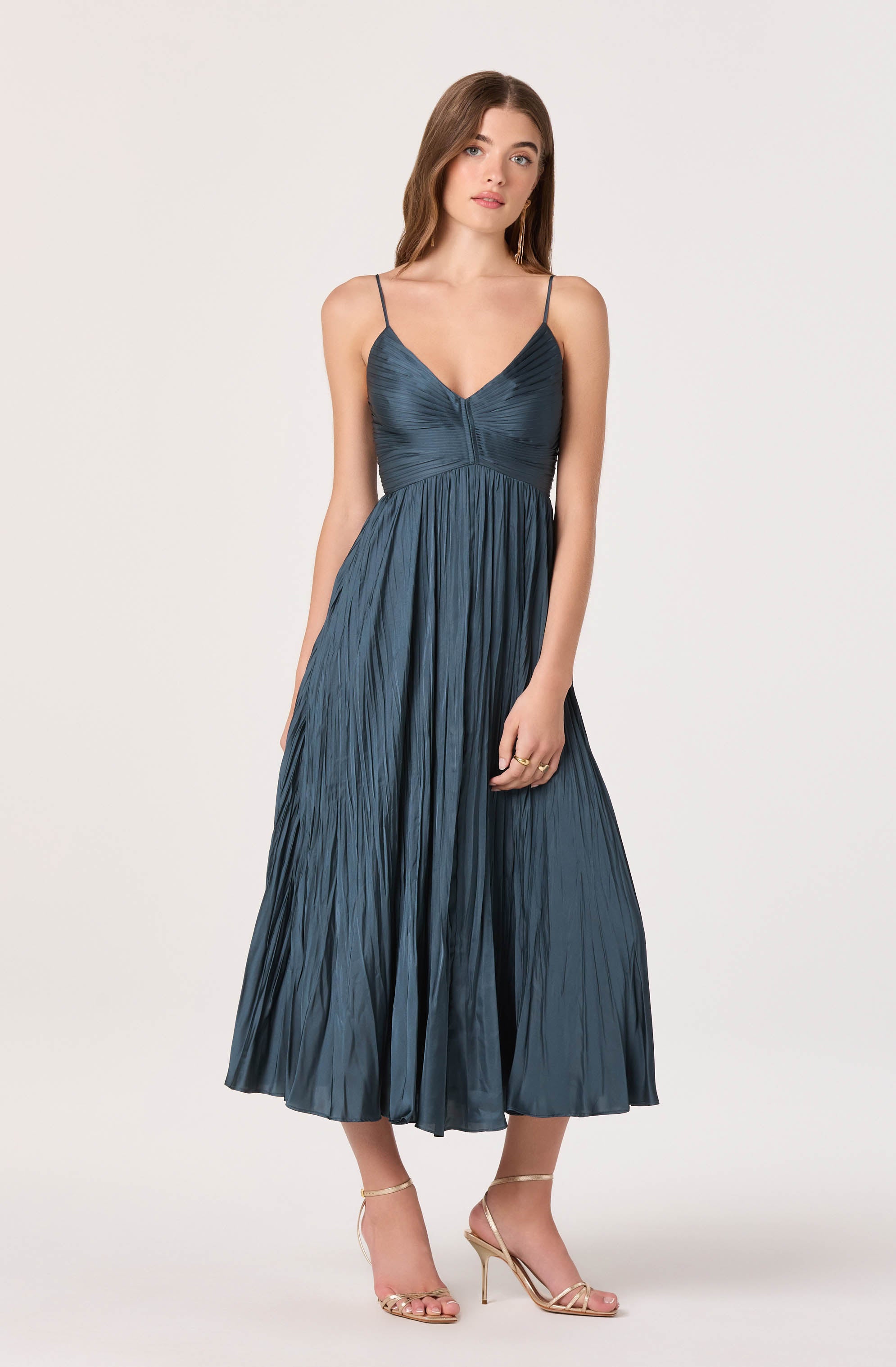A woman is wearing a long, dark blue pleated dress with thin spaghetti straps. The dress has a fitted bodice with a V-neckline and a gathered detail at the bust. The skirt flows in pleats down to her ankles. She is also wearing nude high-heeled sandals with thin straps.