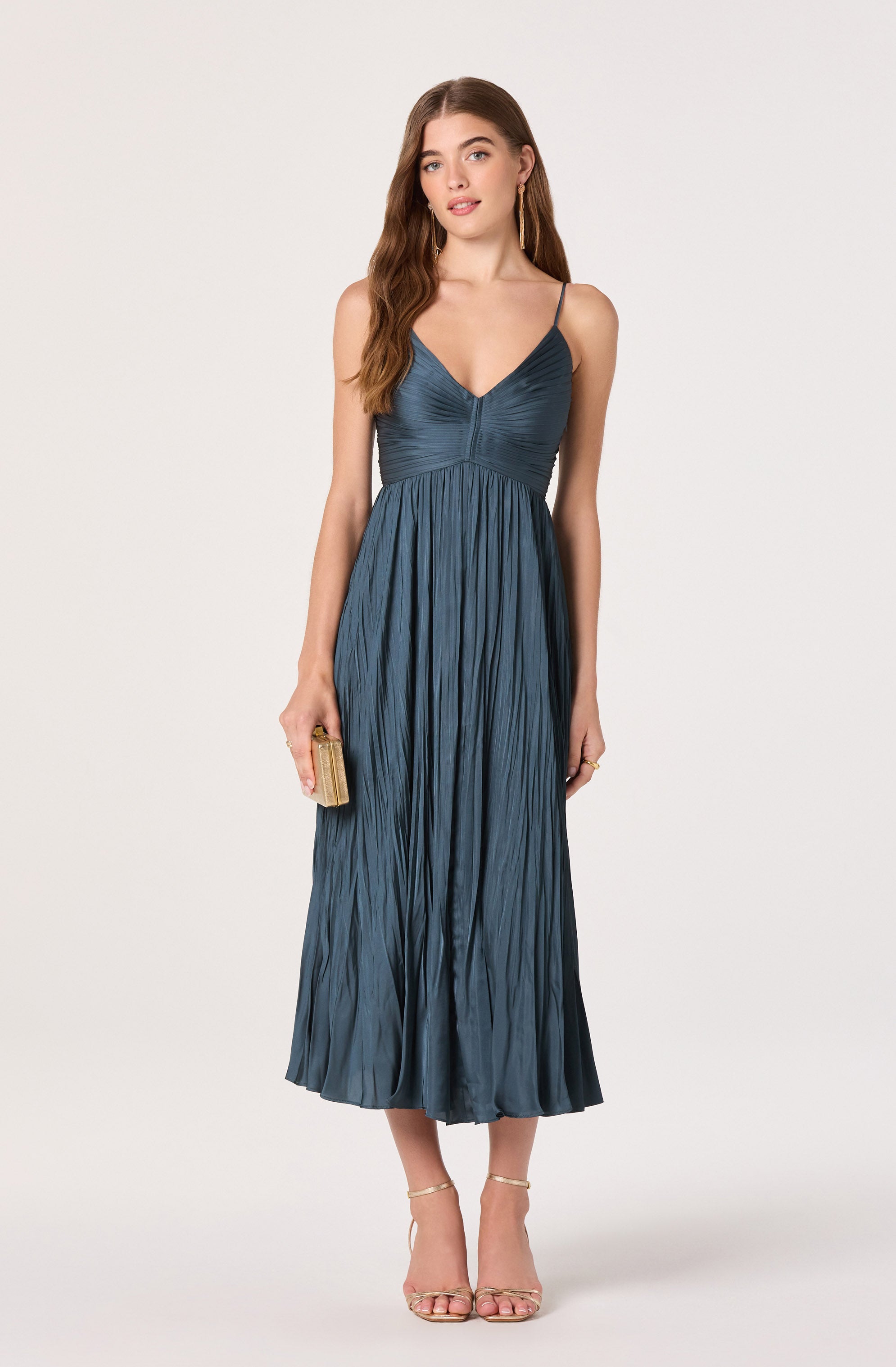 A woman is wearing a long, silky, pleated dress in a deep blue color. The dress has a fitted bodice with thin spaghetti straps and a V-neckline. She is holding a small, gold clutch purse in her right hand and is wearing open-toe high heel sandals.