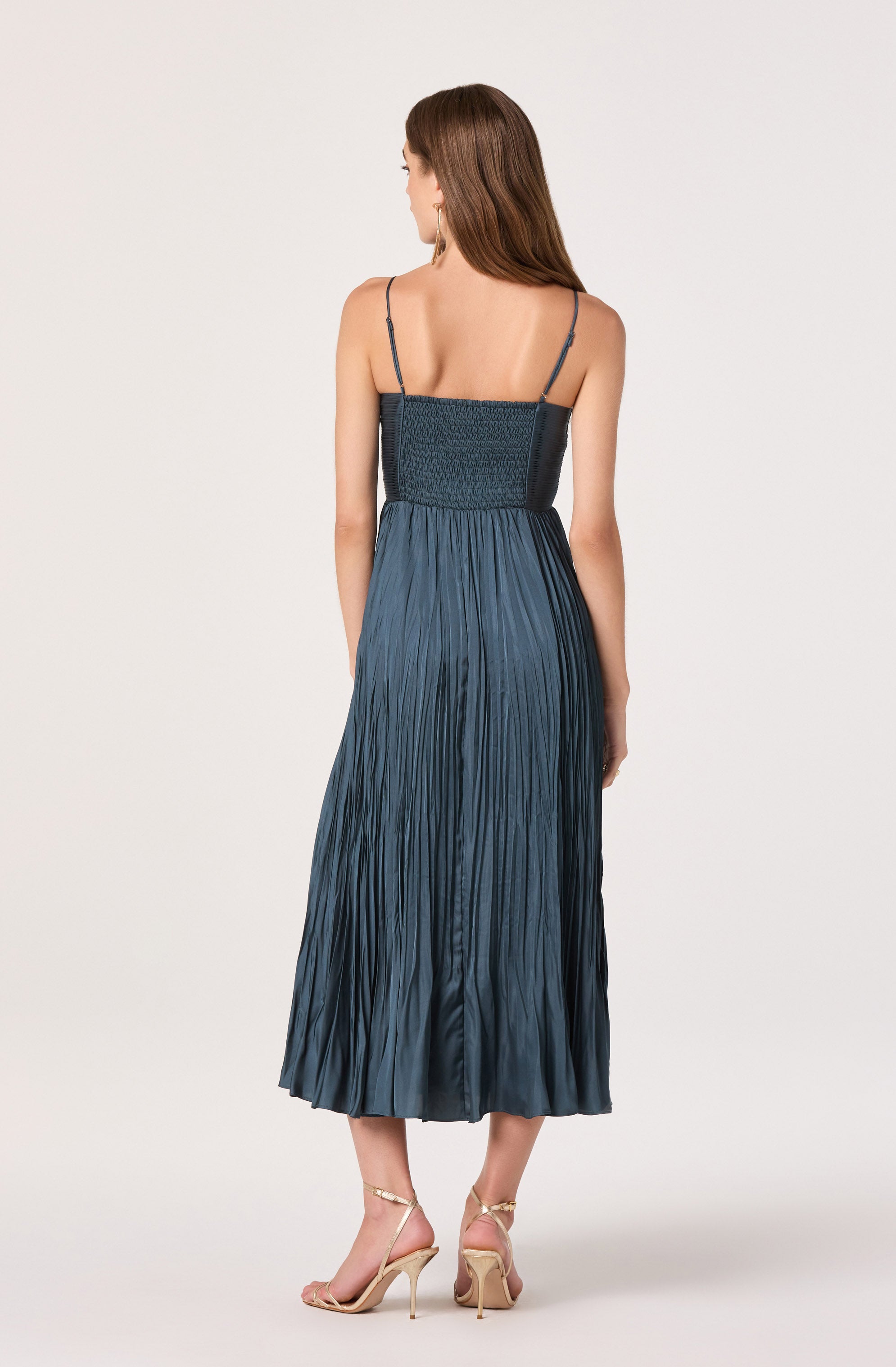 A woman is wearing a dark blue, pleated, sleeveless dress with thin spaghetti straps. The dress has a fitted, smocked bodice and flows down to a midi length. She is also wearing beige high-heeled sandals with thin ankle straps.