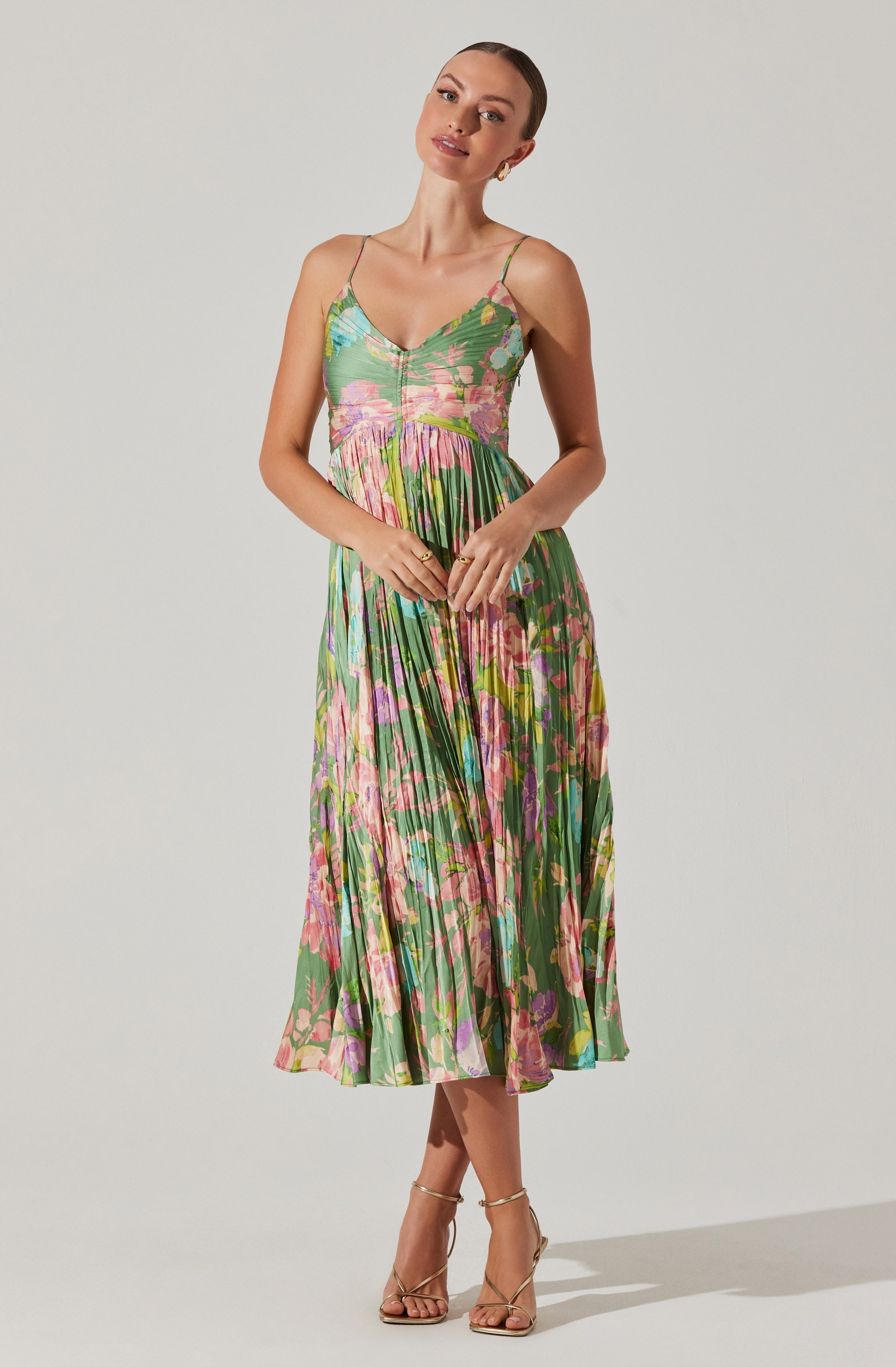 A woman is wearing a sleeveless, V-neck, spaghetti strap dress with a vibrant, multicolored floral pattern. The dress features a fitted bodice and a pleated skirt that falls below the knees. She is also wearing strappy high-heeled sandals.