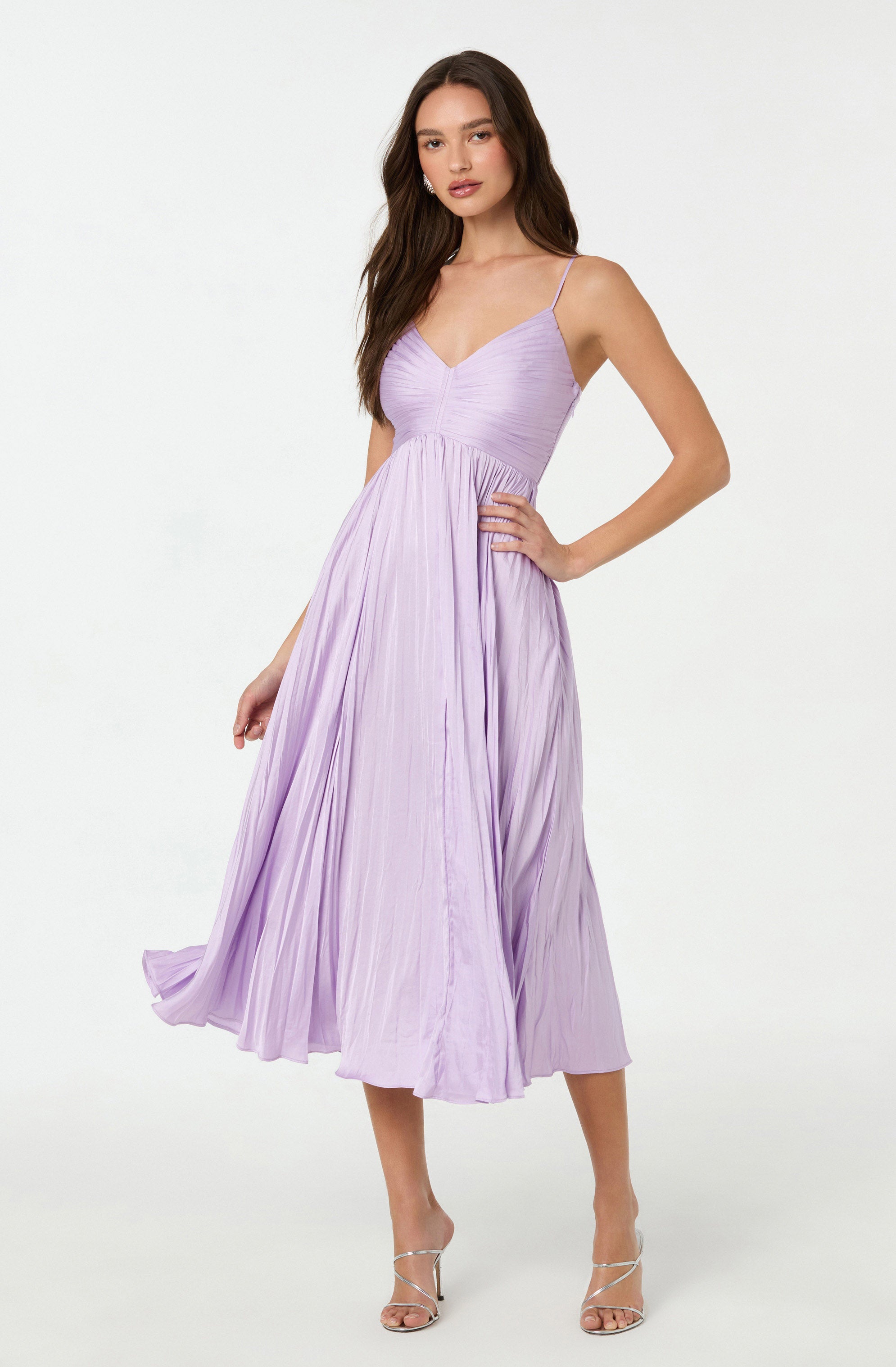 A woman is wearing a lavender-colored dress with spaghetti straps, featuring a fitted, ruched bodice and a flowing, pleated skirt that falls below her knees. She pairs the dress with light-colored, strappy high-heeled sandals.