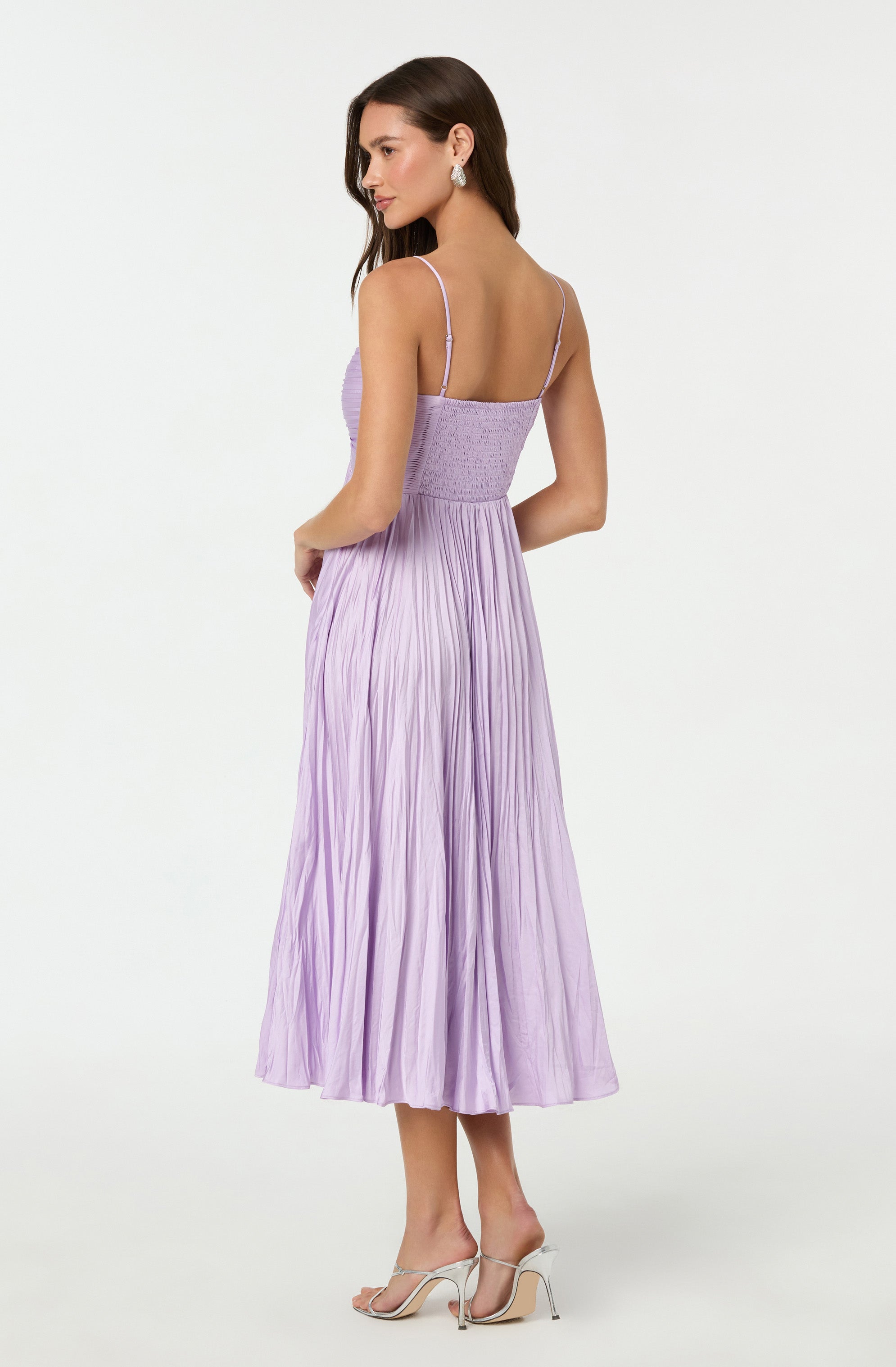 A woman is wearing a light purple, sleeveless dress with spaghetti straps and a fitted, ruched bodice. The skirt is long, flowing, and pleated, reaching mid-calf. She is also wearing white high-heeled sandals.
