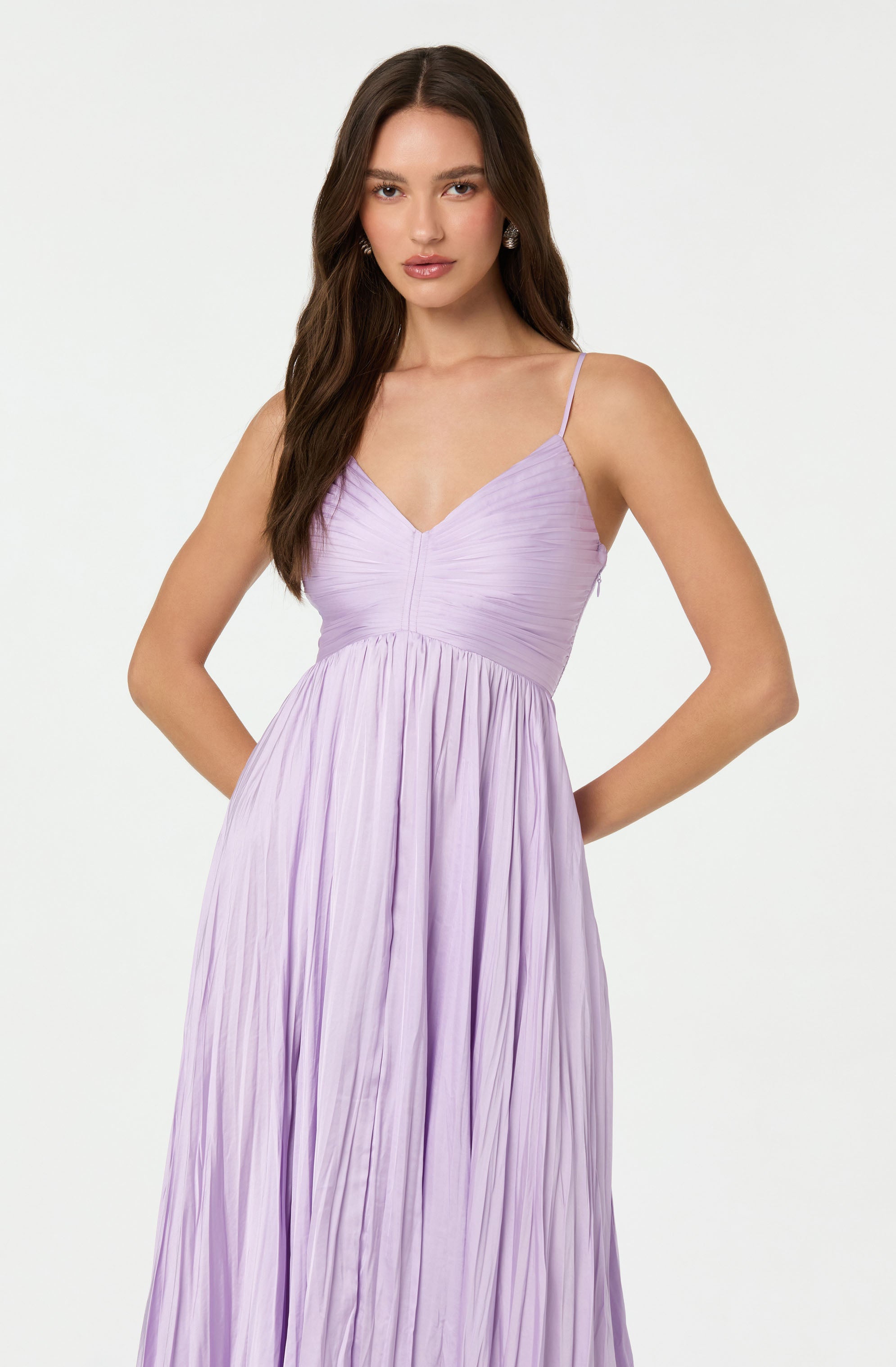 A woman is wearing a light purple, pleated dress with thin spaghetti straps. The dress features a fitted bodice with a gathered, ruched design that extends from the bust to the waist, flowing into a long, flowing skirt with vertical pleats.