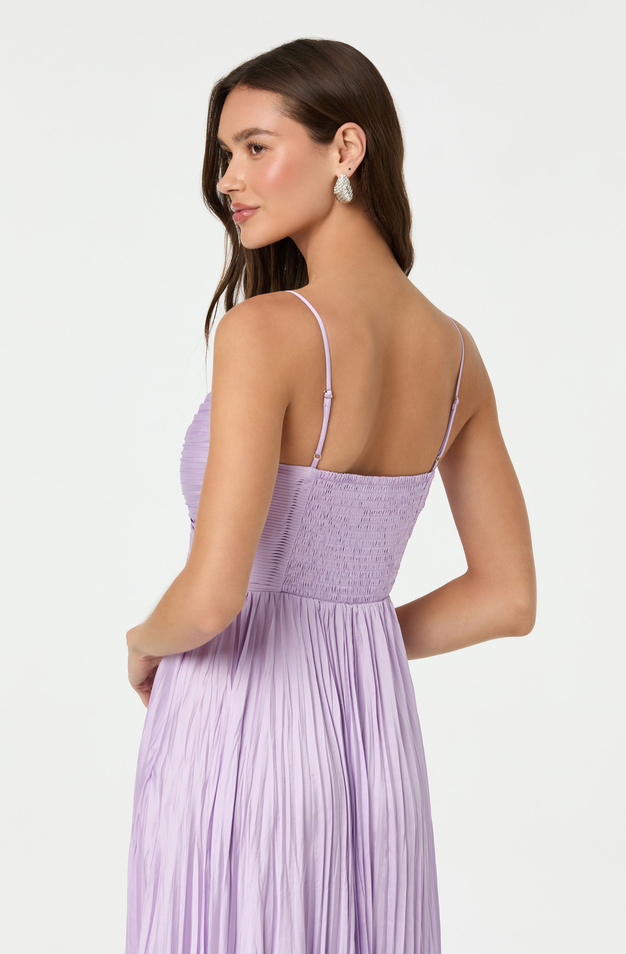 A woman is wearing a light purple, sleeveless dress with thin spaghetti straps. The top portion of the dress features a fitted, smocked bodice with a textured, ruched design. The skirt of the dress is pleated and flows downward in soft, vertical folds. She is also wearing large, ornate earrings.