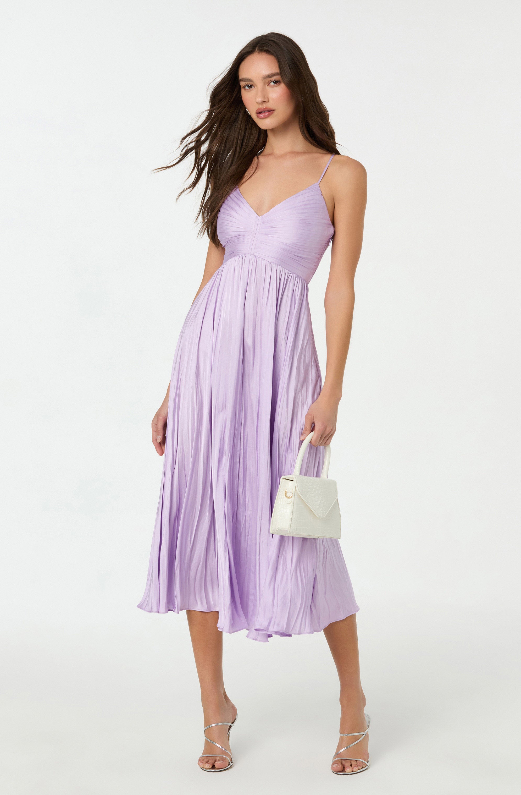 A woman is wearing a light purple, pleated dress with thin spaghetti straps. The dress has a fitted bodice with a V neckline and flows down to mid-calf length. She is holding a small, white handbag and is wearing strappy high-heeled sandals.