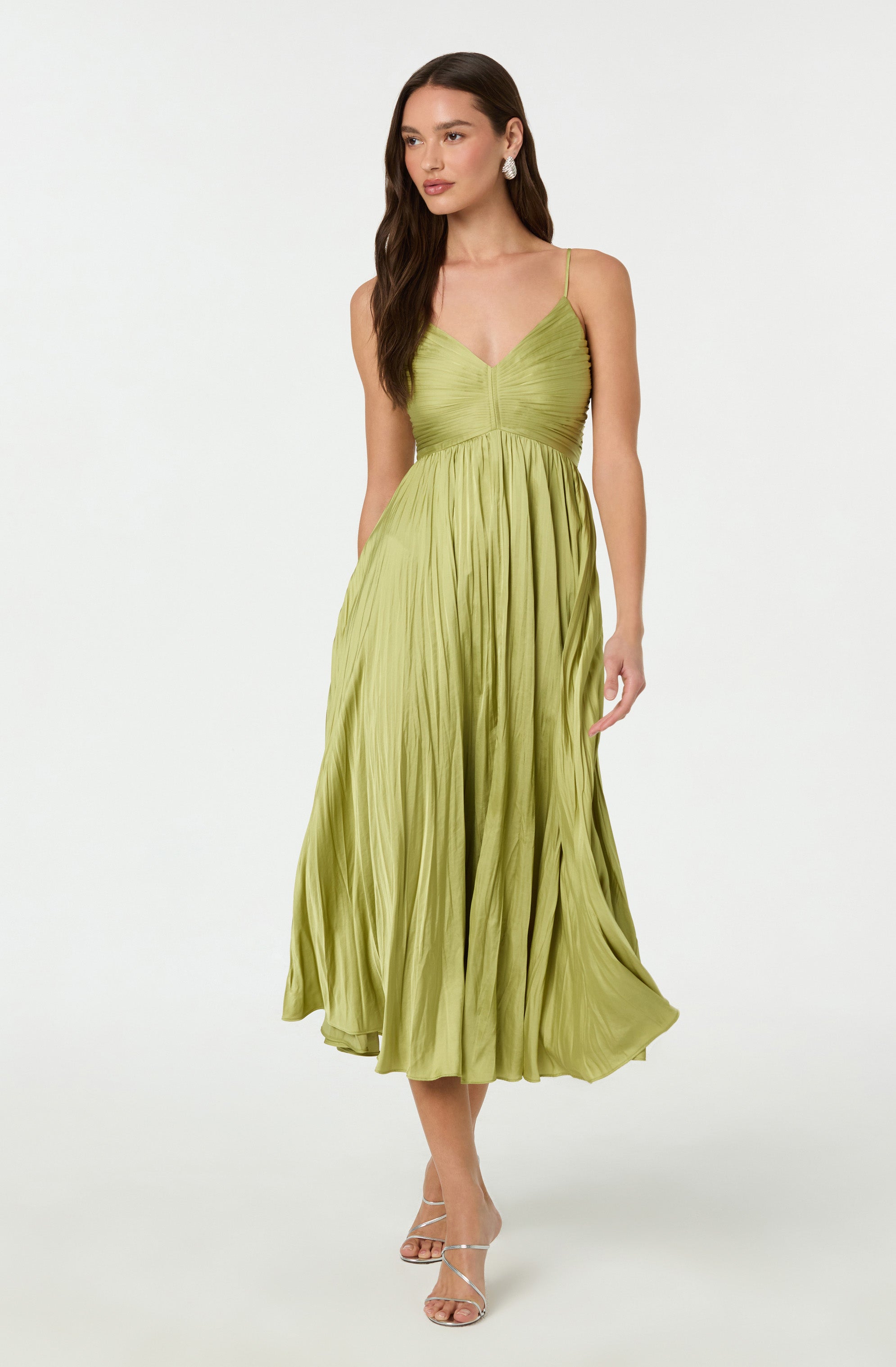 A woman is wearing a light green, pleated midi dress with spaghetti straps and a fitted bodice. She has long, dark hair and is accessorized with dangling earrings. She is also wearing white, high-heeled sandals.