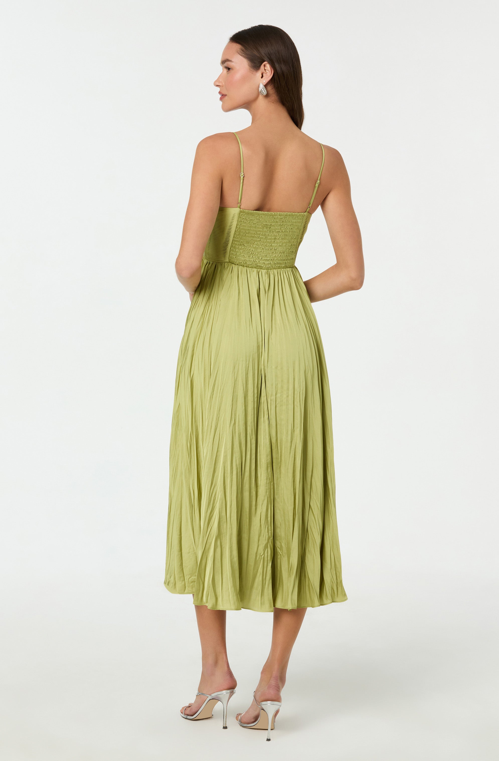 A woman is wearing a light green, sleeveless dress with thin spaghetti straps. The bodice is fitted and has a gathered or shirred detail around the waist. The skirt is long, flowing, and has a crinkled texture with vertical pleats, reaching mid-calf. She is also wearing light-colored high-heeled sandals.