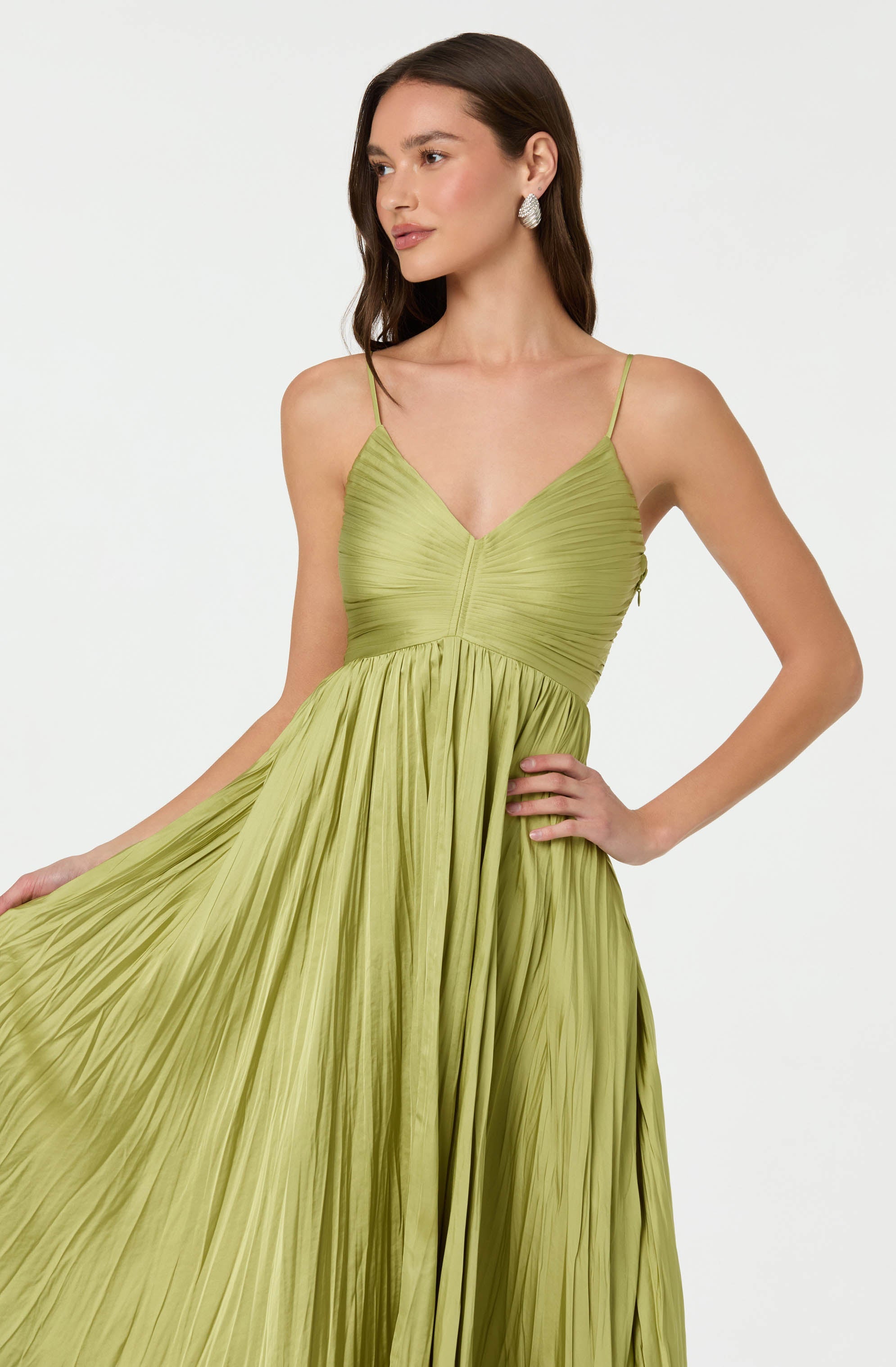 A woman is wearing a light green, pleated dress with thin spaghetti straps. The dress features a fitted bodice with a V neckline and a full, flowing skirt. She accessorizes with large, round earrings.