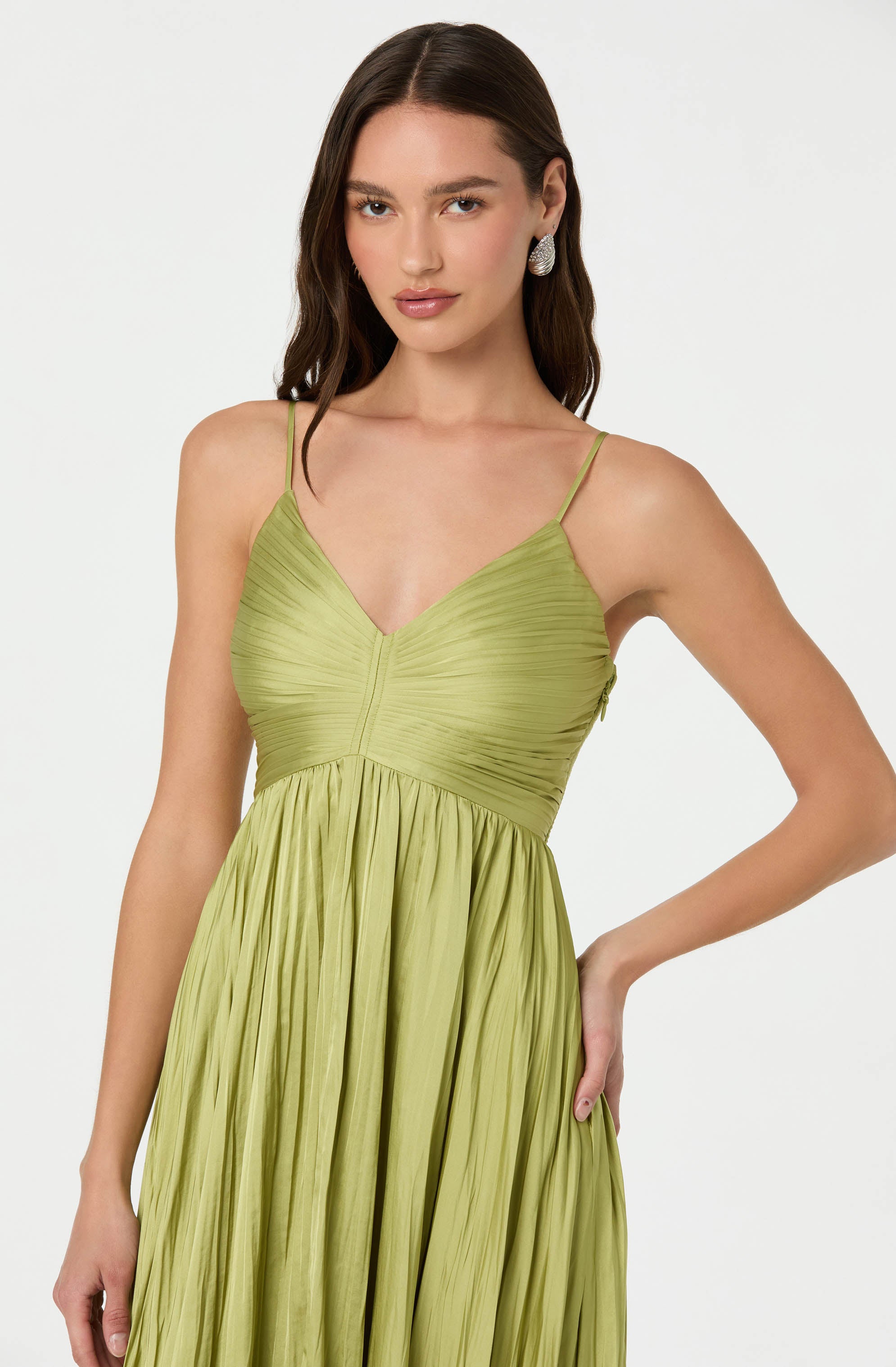 A woman is wearing a light green dress with a pleated, textured fabric. The dress features thin spaghetti straps, a fitted bodice with a V-neckline, and a flowing, gathered skirt. She is accessorized with large, circular earrings.