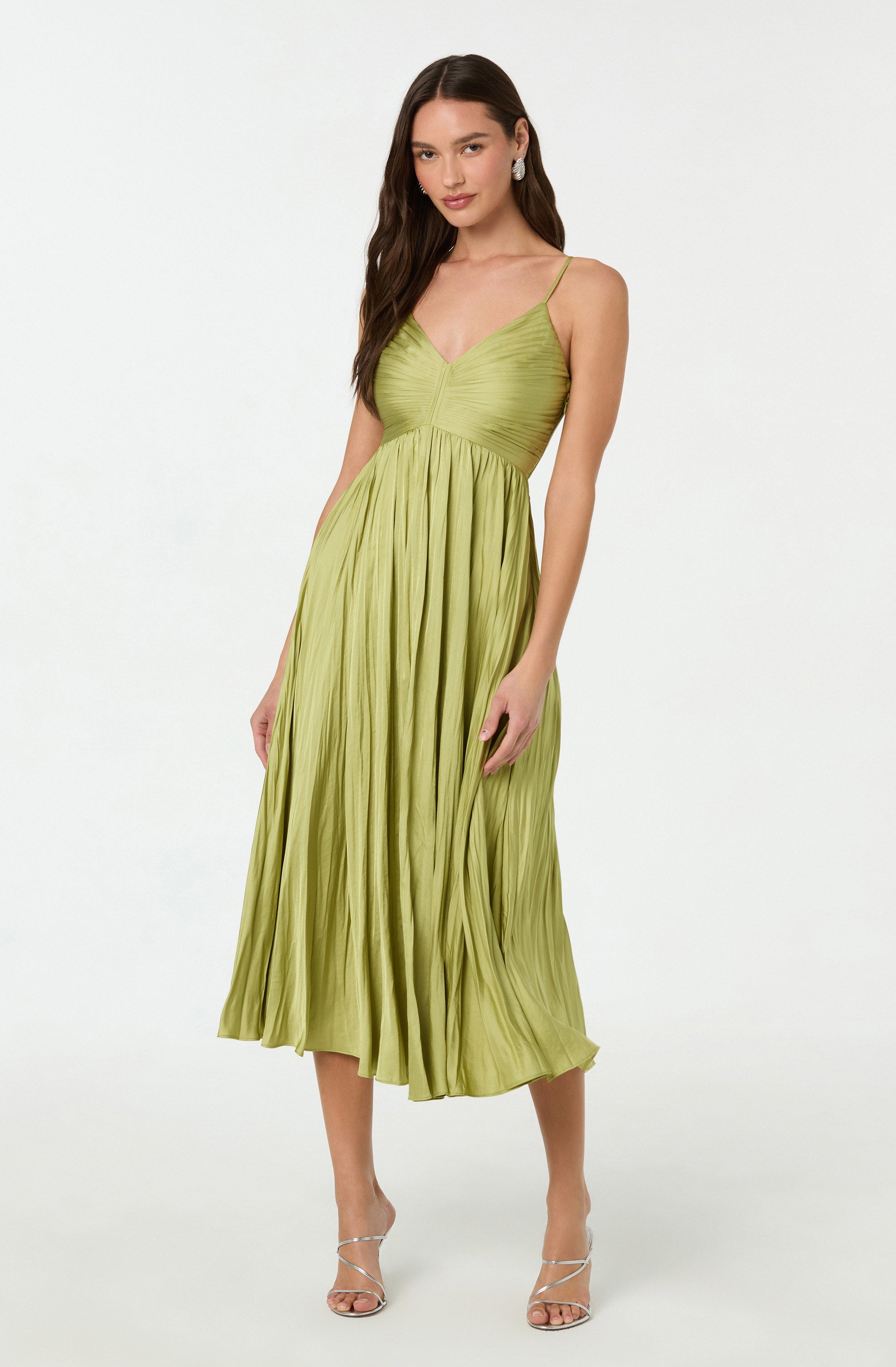 A woman is wearing a light green, pleated, midi-length dress with a fitted bodice, thin spaghetti straps, and a V-neckline. She is also wearing white high-heeled sandals and pearl earrings.
