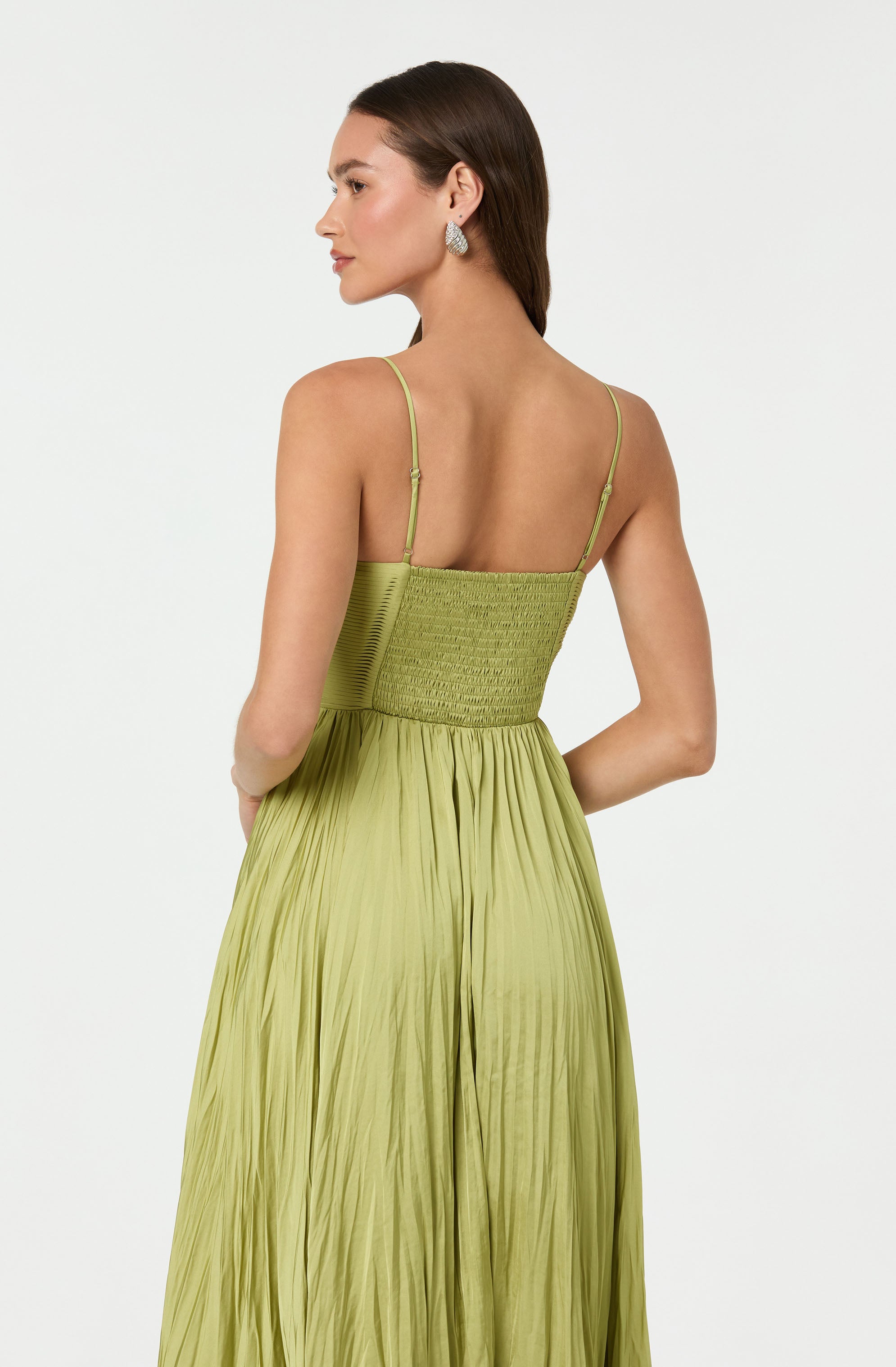 A woman is wearing a light green, pleated dress with thin spaghetti straps. The top part of the dress features a smocked, textured bodice, while the skirt is flowing with vertical pleats. The dress has a gradient effect, with the color lighter at the top and darker at the bottom.