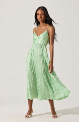 A woman is wearing a sleeveless, light green midi dress with thin spaghetti straps. The dress features a fitted bodice with a V-neckline and a gathered waist, flowing into a pleated skirt. The fabric has a polka dot pattern with white dots scattered throughout. She is also wearing white open-toed high-heeled sandals.
