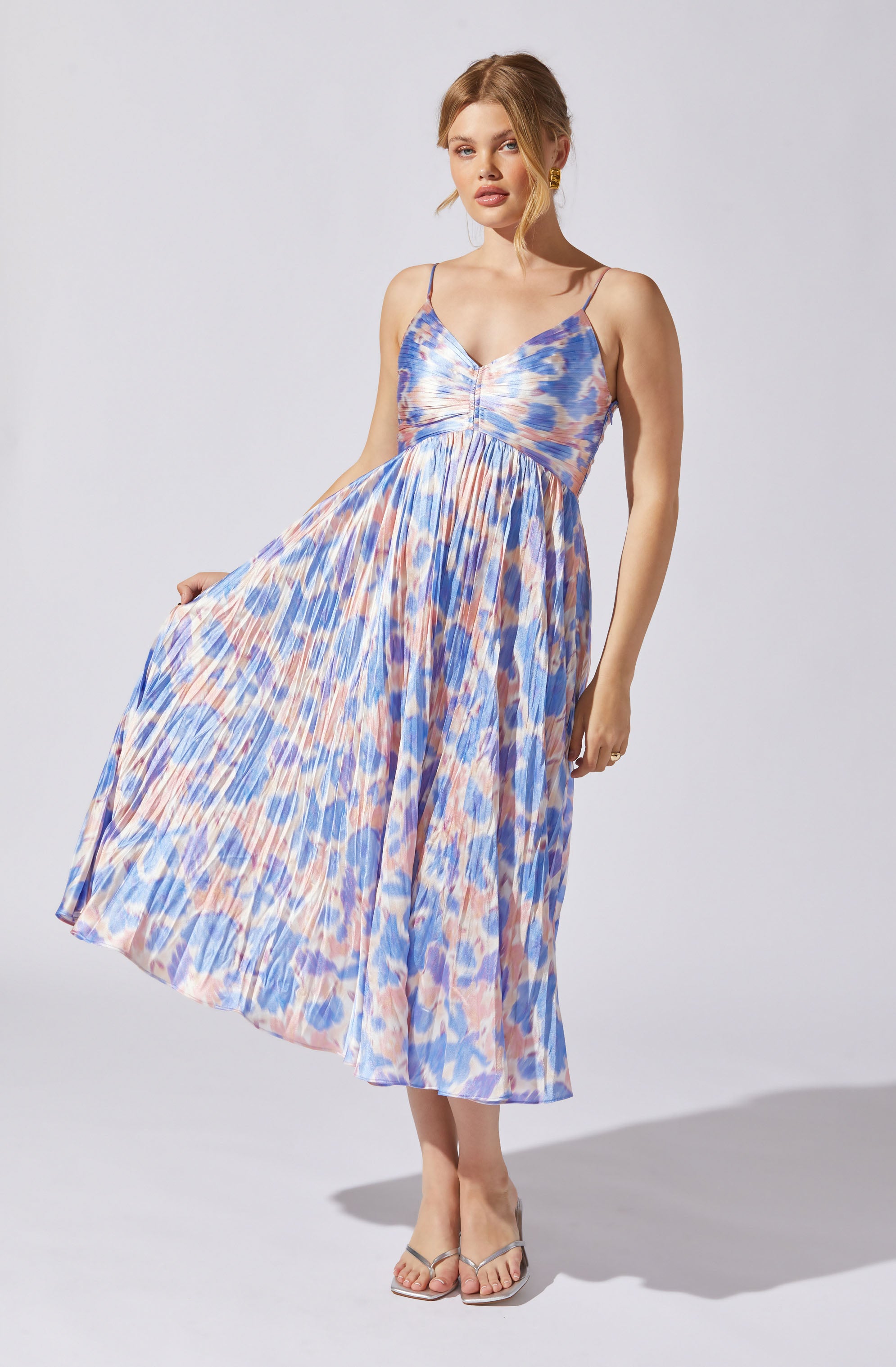 A woman is wearing a sleeveless, V-neck dress with thin spaghetti straps. The dress has a colorful, abstract pattern in shades of blue, peach, and purple, with a fitted bodice and a full, pleated skirt that reaches mid-calf. She is also wearing open-toed high heels.