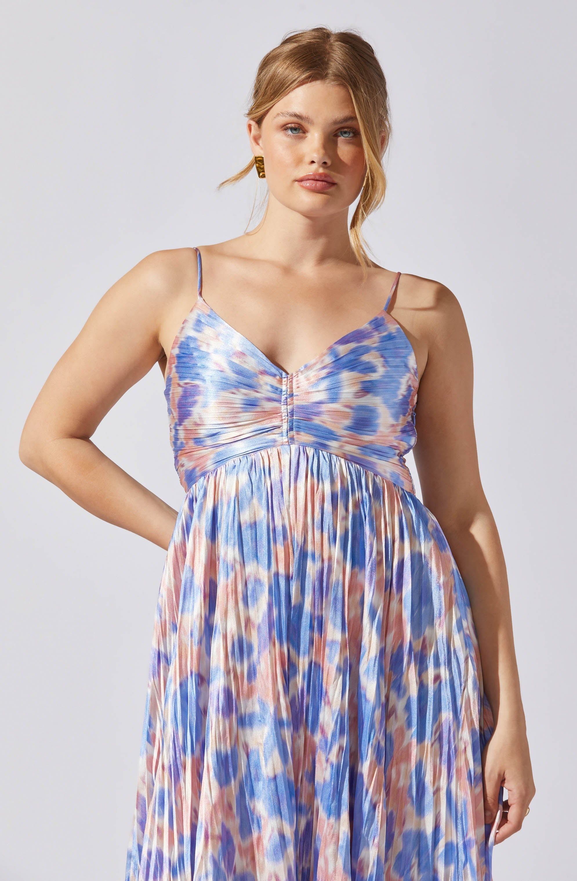 A woman is wearing a sleeveless, flowy dress with thin spaghetti straps. The dress features a ruched bodice and an empire waistline, with a colorful, abstract watercolor pattern in shades of blue, pink, and cream.