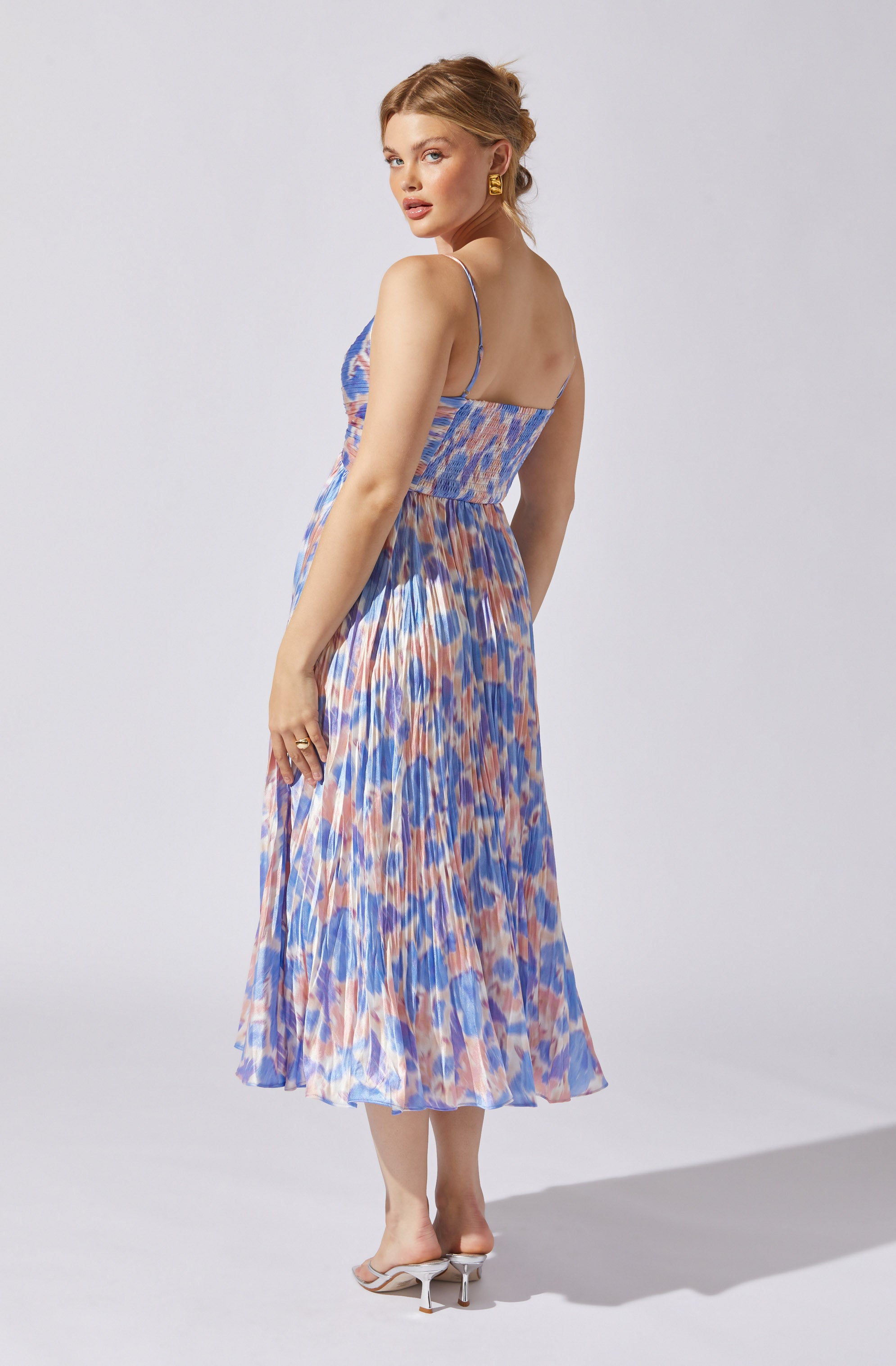 A woman is wearing a sleeveless, strappy dress with a colorful, abstract pattern in shades of blue, peach, and white. The dress features a fitted bodice with a smocked waist and a flowing, pleated midi-length skirt. She is also wearing open-toe heels with a clear strap and a small heel.