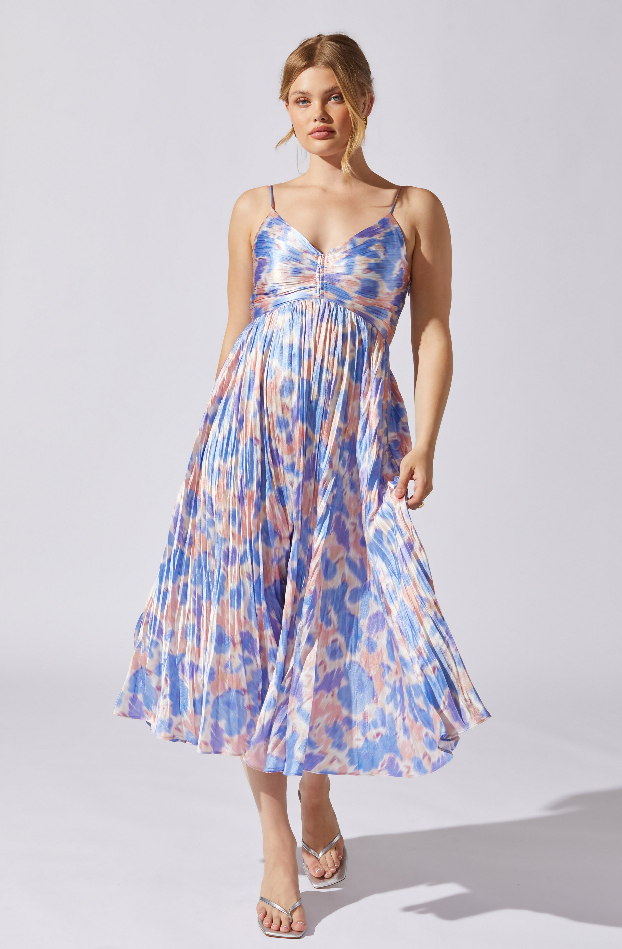 A woman is wearing a sleeveless, spaghetti strap maxi dress with a colorful, abstract animal print pattern in shades of blue, pink, and beige. The dress features a fitted bodice with a gathered detail at the center and a flowing, pleated skirt. She is also wearing clear high-heeled sandals.