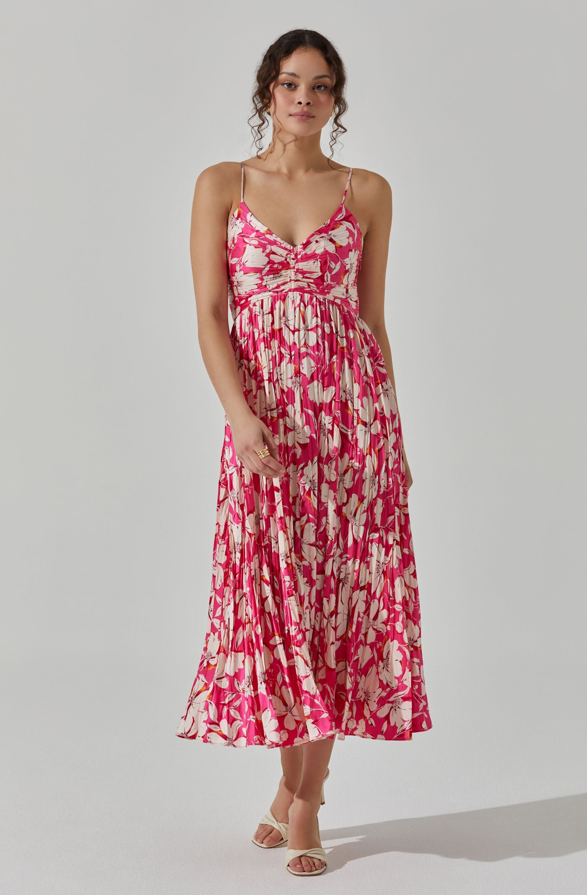 A woman is wearing a sleeveless, pink and beige floral-printed dress with thin spaghetti straps and a V-neckline. The dress has a fitted bodice with a gathered detail at the bust and a flowing, pleated skirt that reaches mid-calf. She is also wearing open-toed, beige high-heeled sandals.