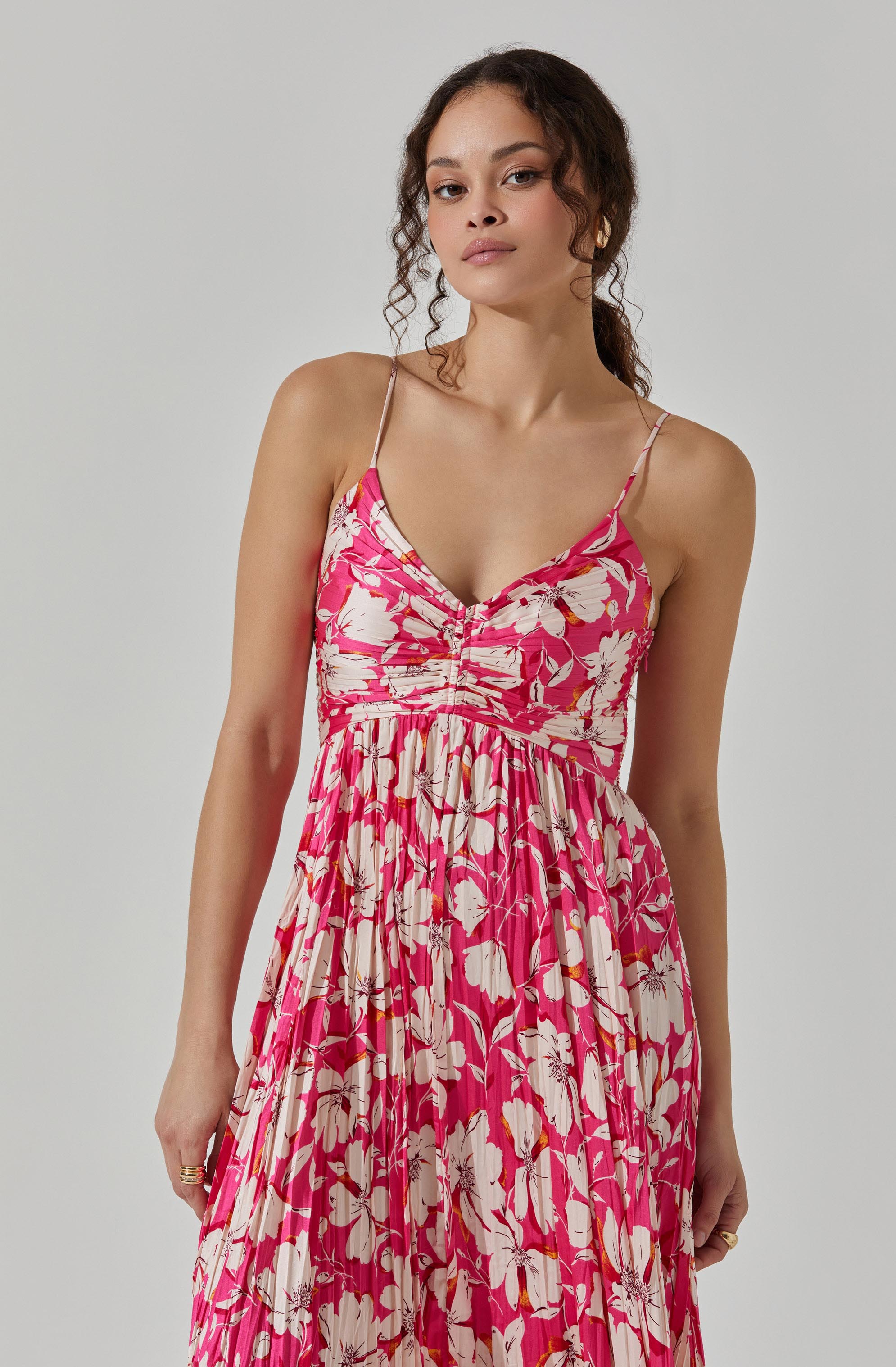 A woman is wearing a sleeveless, pink dress with thin spaghetti straps. The dress features a floral pattern with large white and light pink flowers and green leaves. It has a fitted bodice with ruching detail and a flowing, pleated skirt.