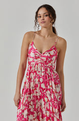 A woman is wearing a sleeveless, pink dress with thin spaghetti straps. The dress features a floral pattern with large white and light pink flowers and green leaves. It has a fitted bodice with ruching detail and a flowing, pleated skirt.
