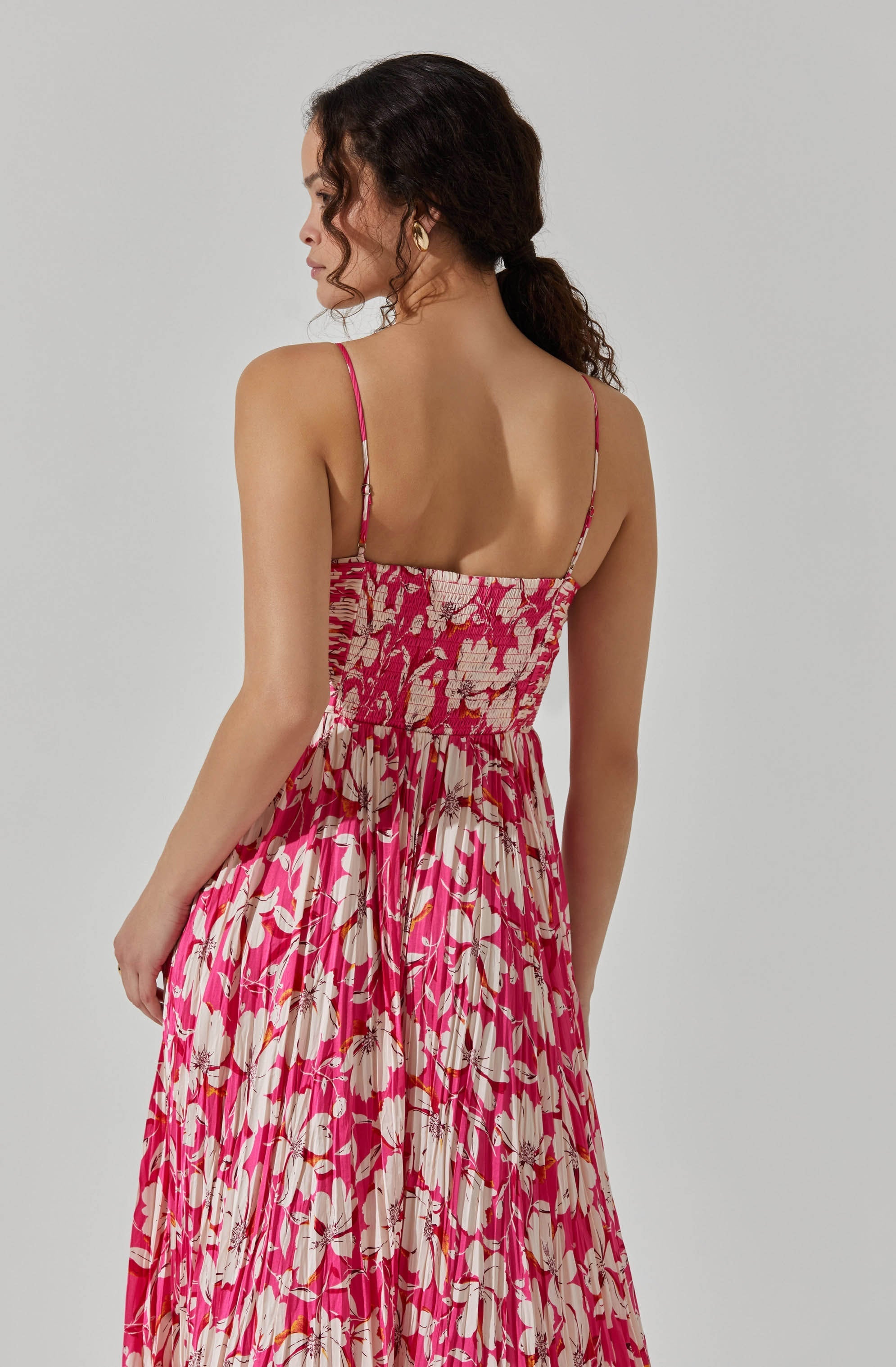 A woman is wearing a sleeveless, spaghetti-strap dress with a fitted, smocked bodice and a flowing, pleated skirt. The dress has a vibrant floral pattern with shades of pink, white, and beige.