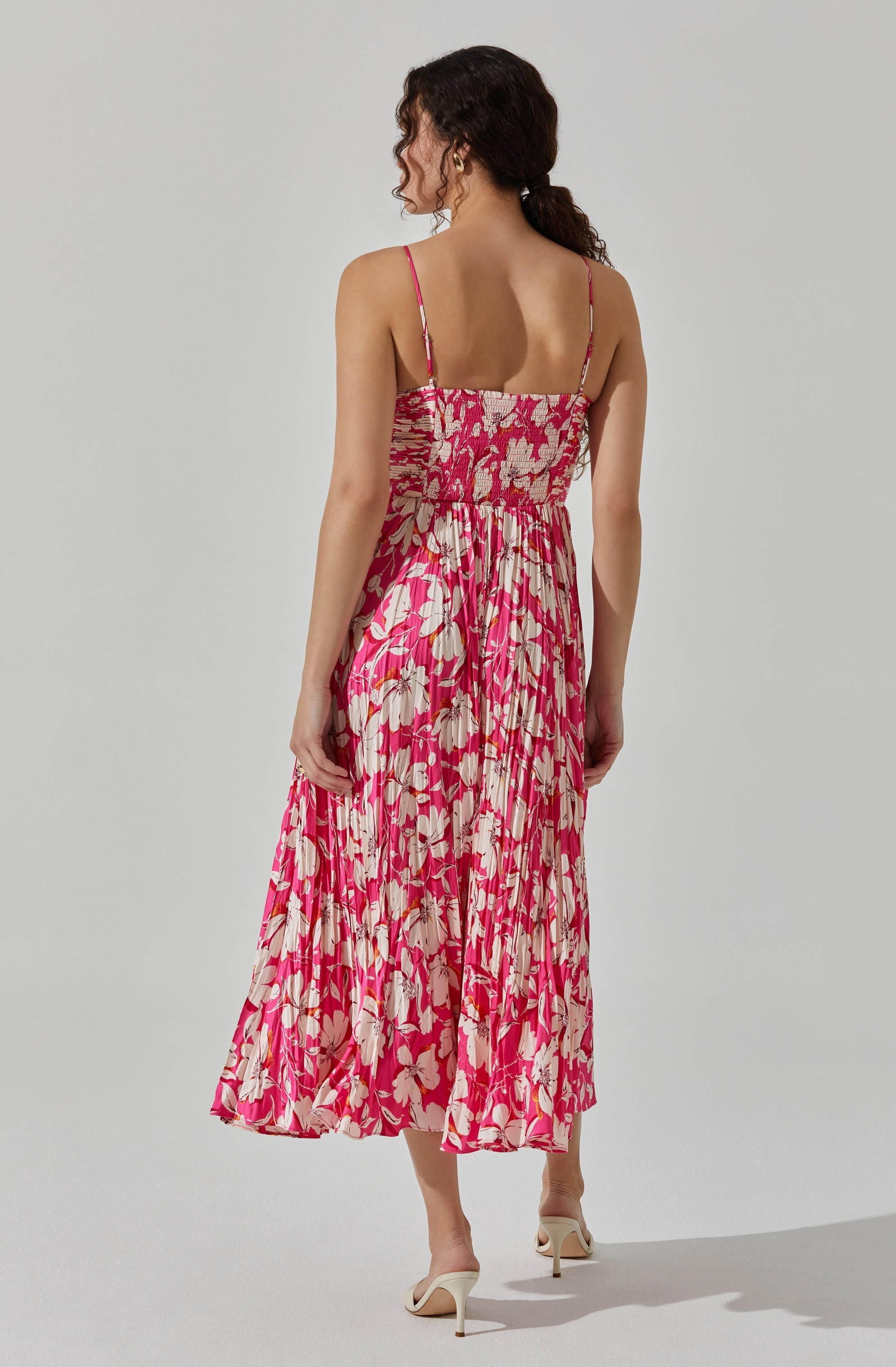 A woman is wearing a sleeveless, ankle-length dress with thin spaghetti straps. The dress features a vibrant pink and beige floral pattern, with a gathered, smocked bodice and a flowing pleated skirt. She is also wearing light-colored, open-toed high heels.
