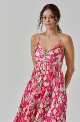 A woman is wearing a sleeveless, pink dress with a floral pattern featuring large white and beige flowers. The dress has thin spaghetti straps and a fitted bodice with a gathered detail at the center. The skirt is long, flowing, and pleated, creating a loose and elegant silhouette.