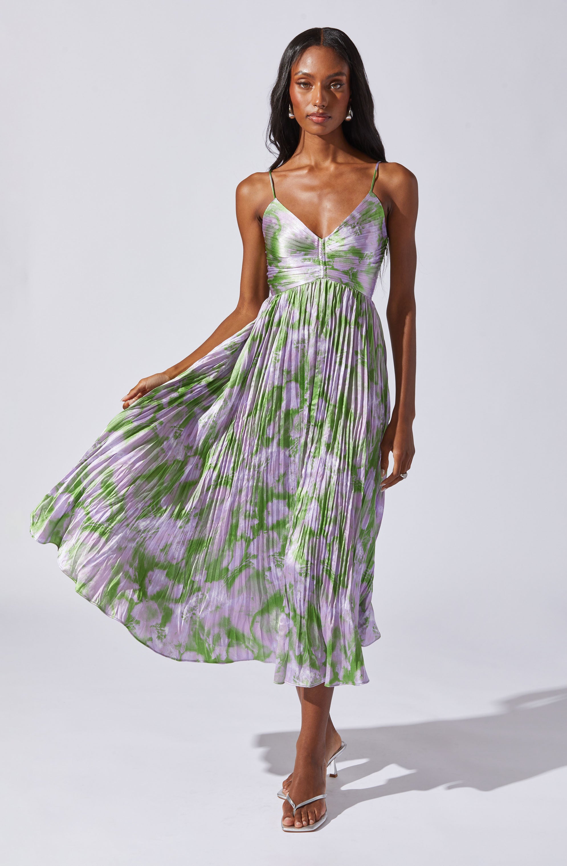 A woman is wearing a sleeveless, V-neck, spaghetti strap dress with a flowing, pleated skirt. The dress features a tie-dye pattern in shades of green and purple. She is also wearing open-toe sandals with a clear strap.