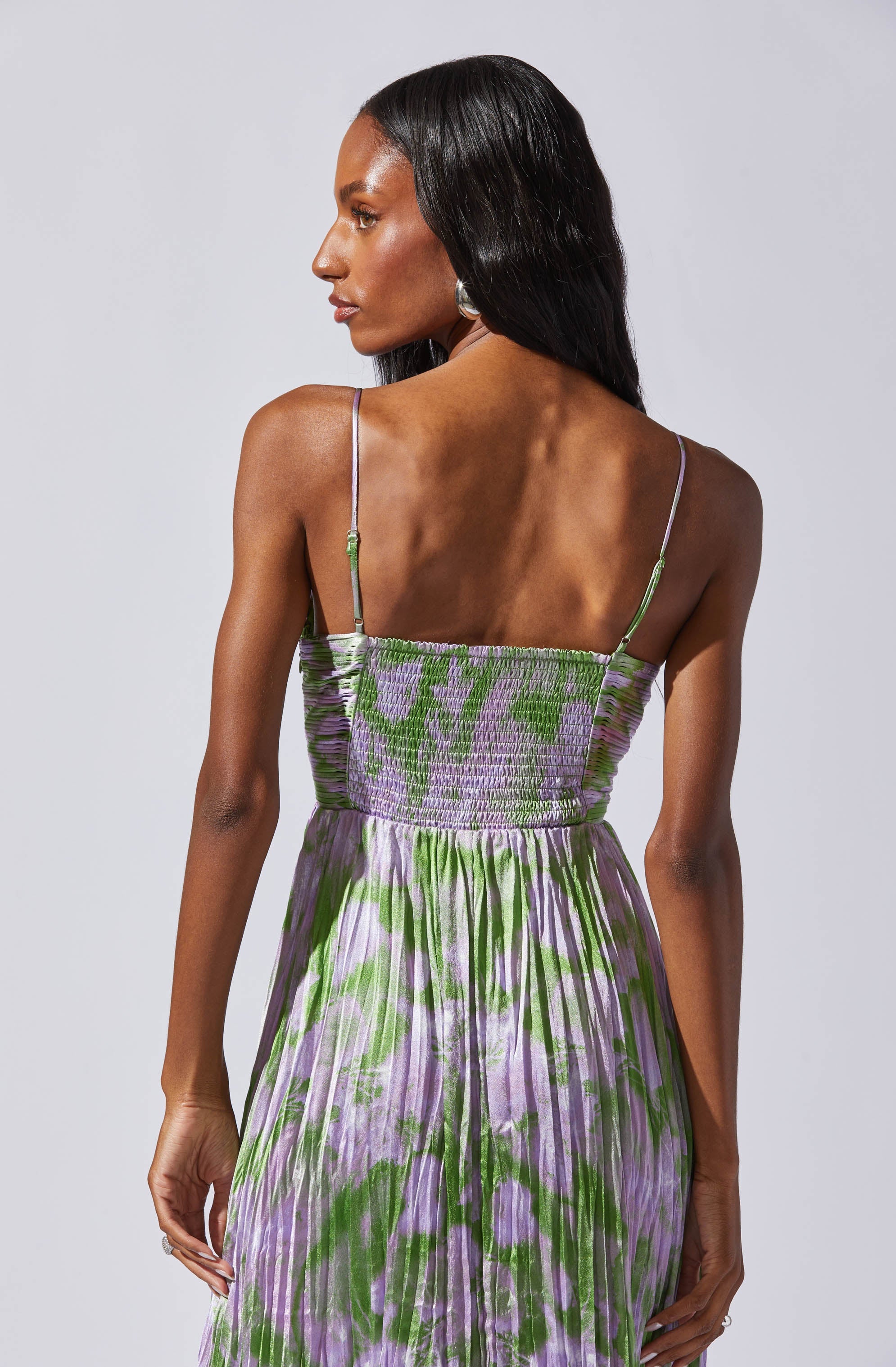 A woman is wearing a sleeveless, floral-patterned dress with thin spaghetti straps. The dress features a fitted, smocked bodice and a flowing skirt with purple and green botanical prints.
