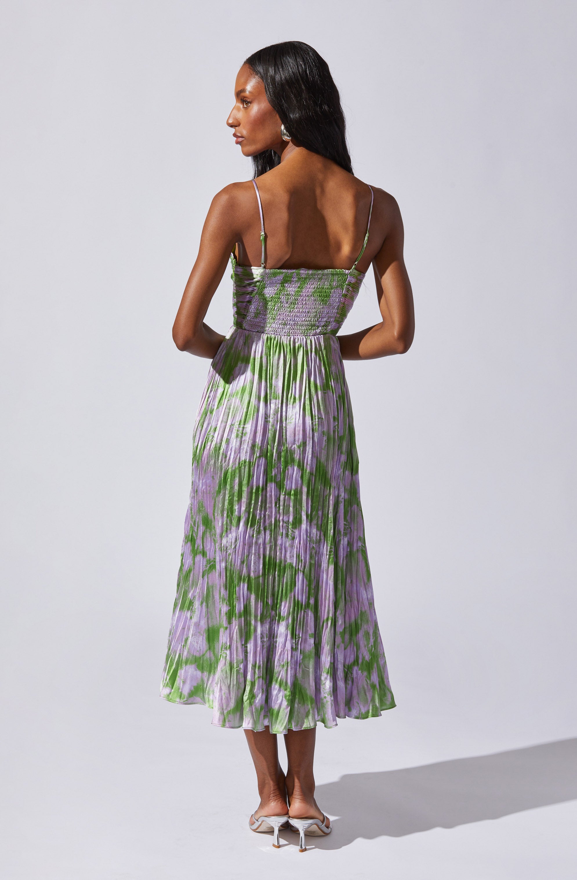 A woman is wearing a sleeveless, ankle-length dress with thin spaghetti straps. The top portion of the dress features a smocked, fitted bodice with a pattern of green and purple colors. The skirt is flowy with a pleated design, showcasing a vibrant green and purple floral or abstract pattern.