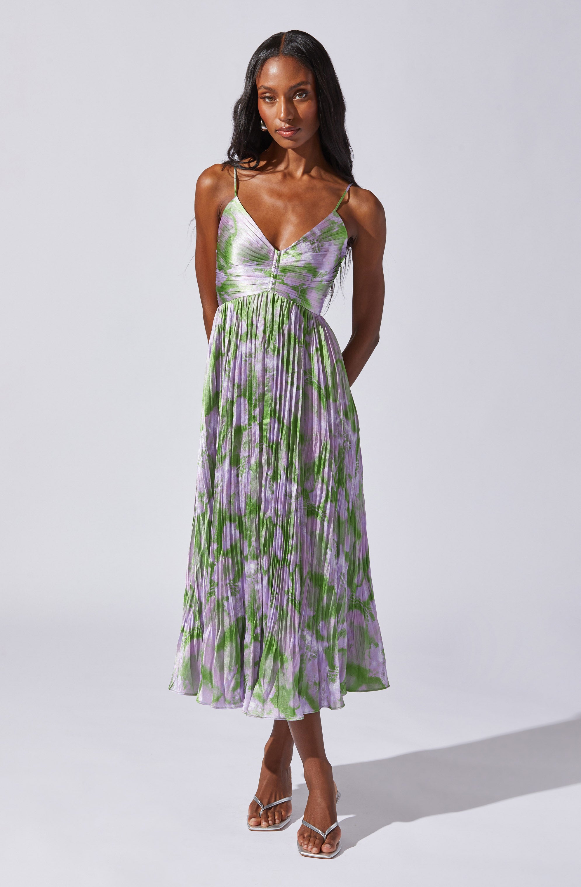 A woman is wearing a sleeveless, V-neck, spaghetti strap dress with a fitted bodice and a flared, pleated skirt that falls below the knees. The dress features a multicolored pattern with shades of purple, green, and white.