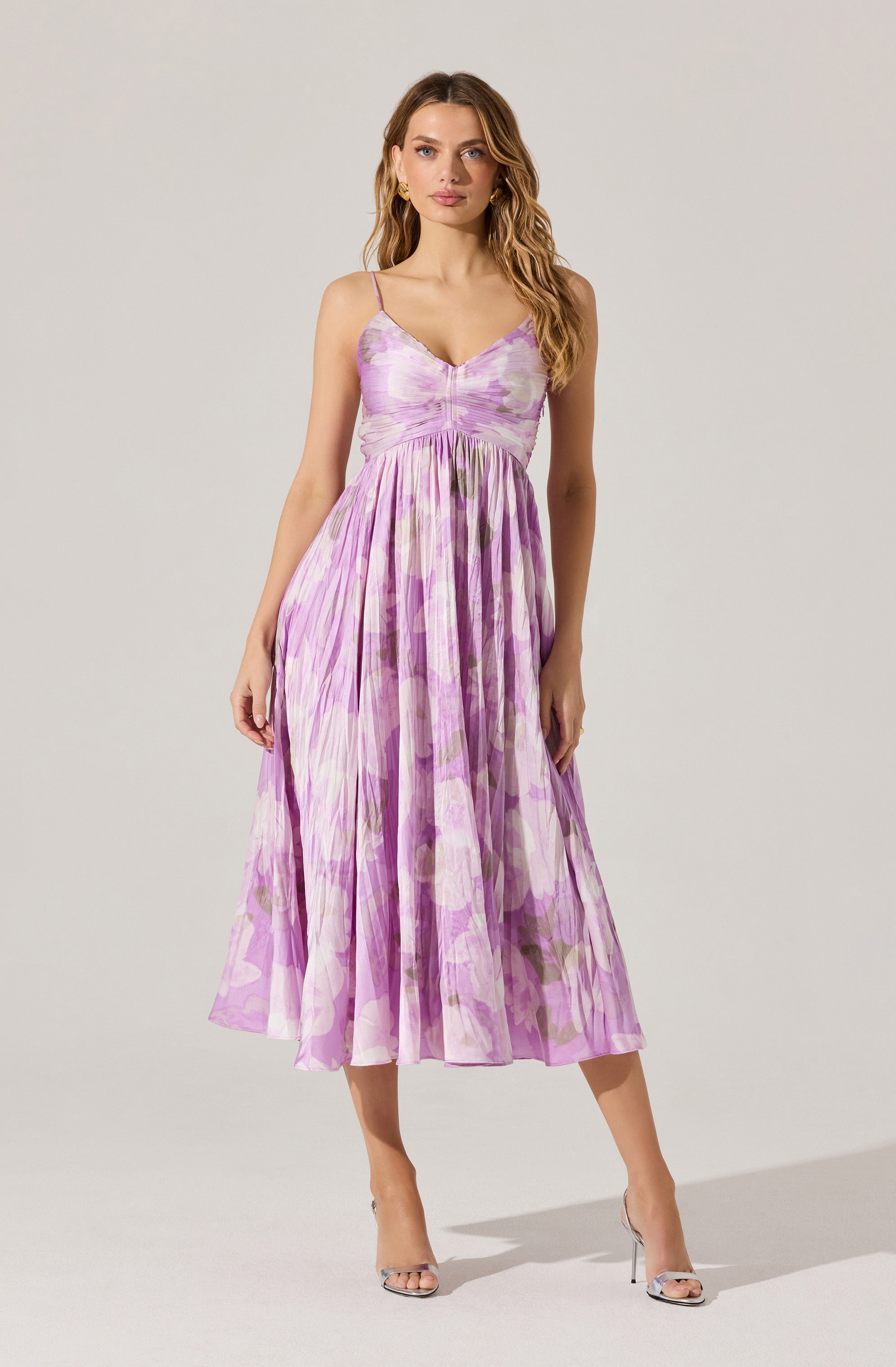 A woman is wearing a sleeveless, midi-length dress with spaghetti straps. The dress has a fitted bodice and a gathered waist, with a flowing skirt. The fabric features a watercolor-like floral pattern in shades of pink, purple, and green. She is also wearing clear high-heeled sandals.