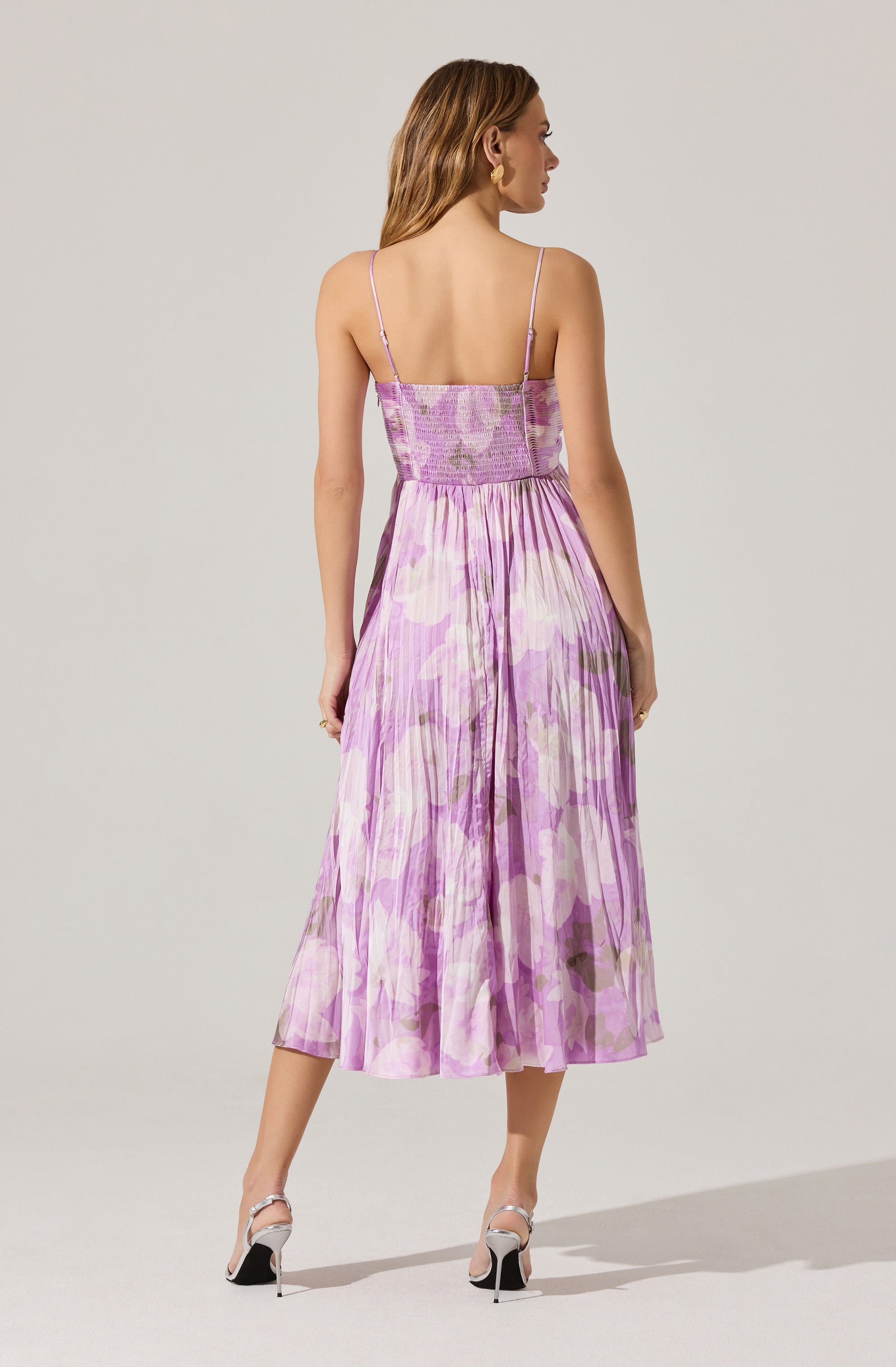 A woman is wearing a sleeveless dress with thin spaghetti straps. The dress features a fitted bodice with shirring at the top and a high-waisted, gathered skirt that falls below the knees. The fabric has a watercolor-like lavender and pink floral pattern with hints of green. She is also wearing high-heeled shoes with ankle straps.