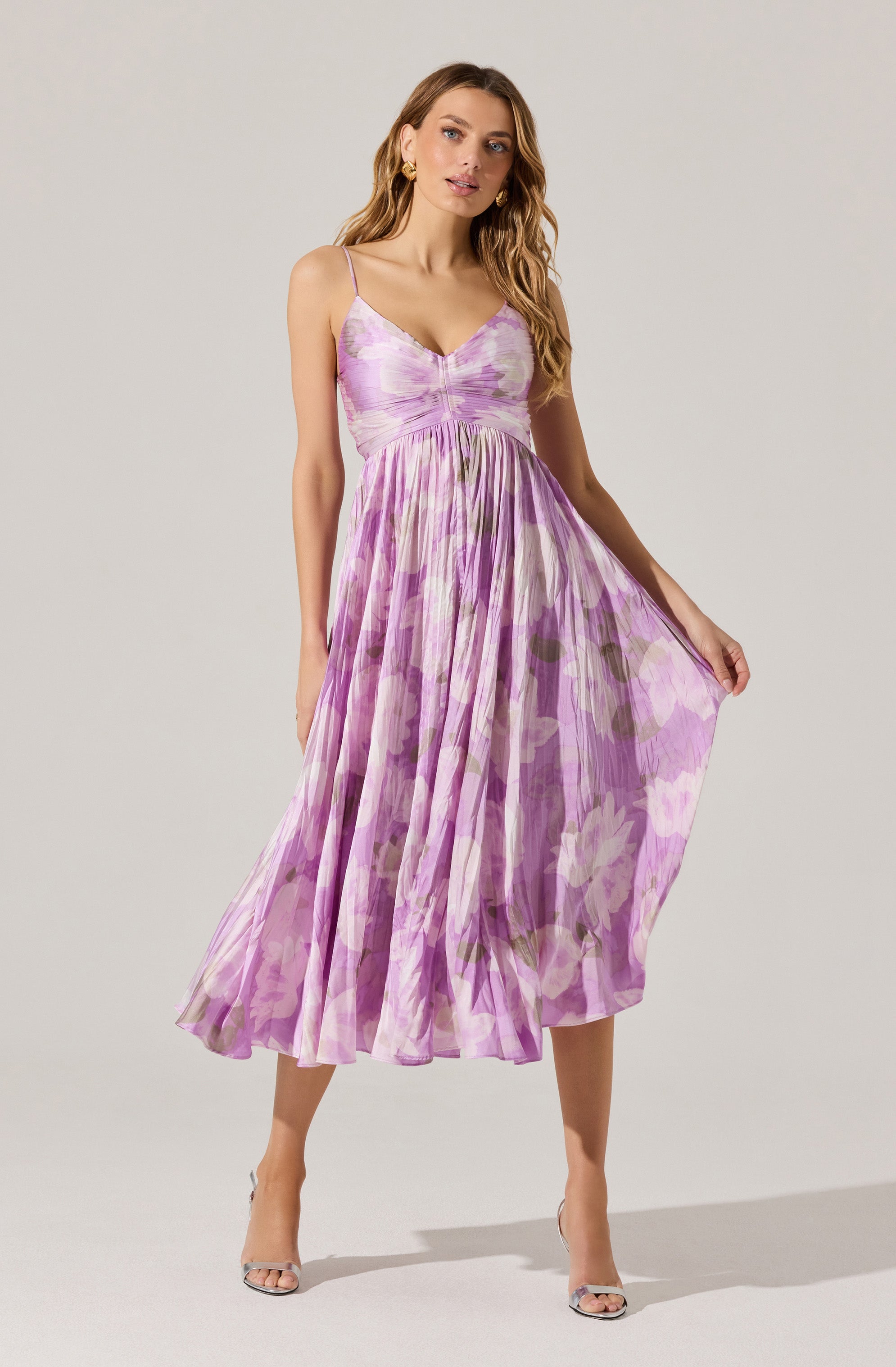 The woman is wearing a sleeveless, flowy midi dress with thin spaghetti straps. The dress features a fitted bodice with subtle ruching at the center and an empire waistline. The fabric has a light, airy quality with a floral pattern in shades of purple, lavender, and hints of green, creating a soft and feminine look. The skirt is pleated, giving it texture and movement. She pairs the dress with open-toe heels.