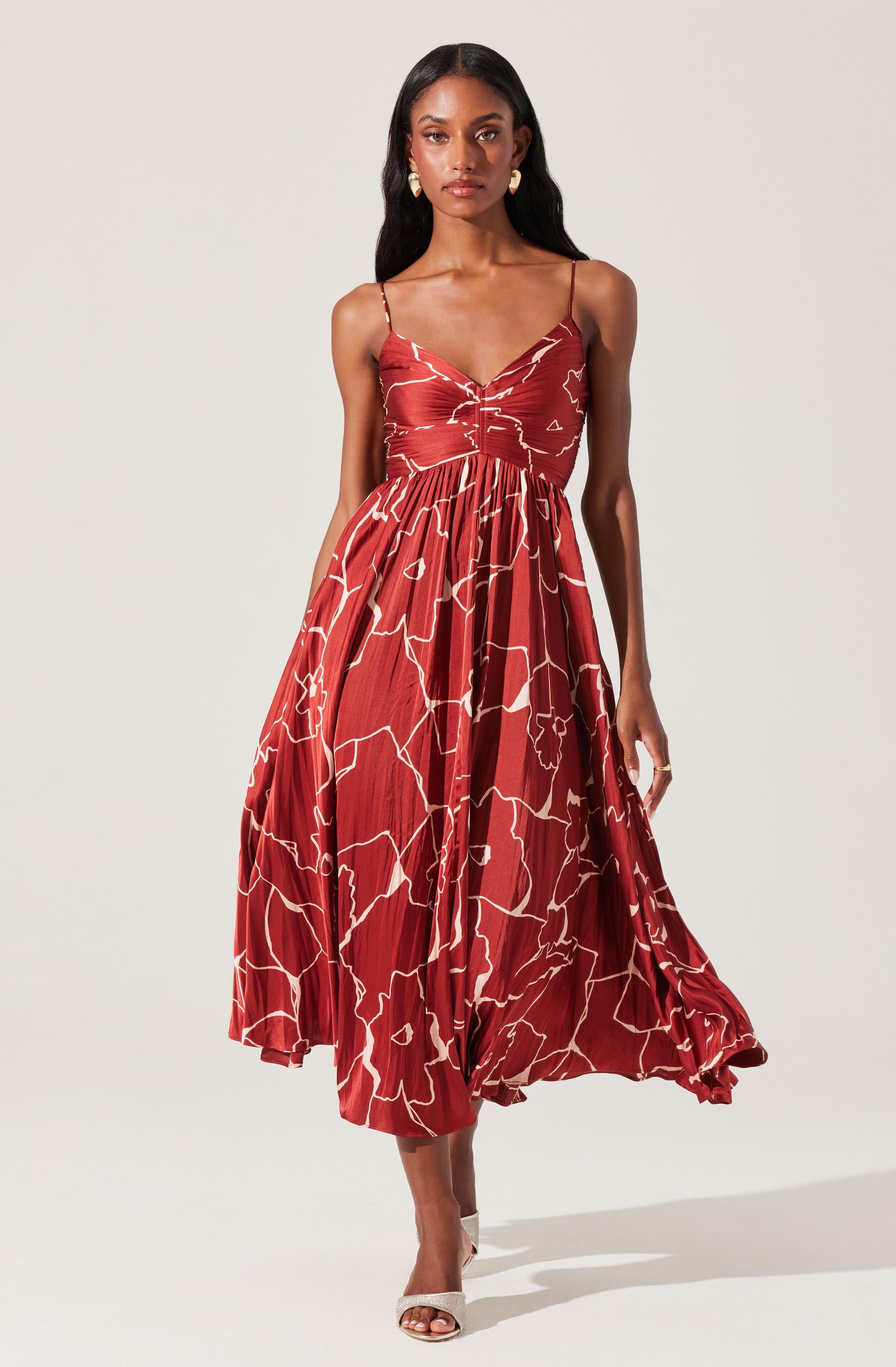 The dress is a sleeveless, ankle-length garment with thin spaghetti straps. It features a fitted bodice with a sweetheart neckline and a gathered waist. The fabric is a rich red color with an abstract, irregular white line pattern throughout. The skirt is flowy and pleated, giving it a voluminous and elegant appearance.