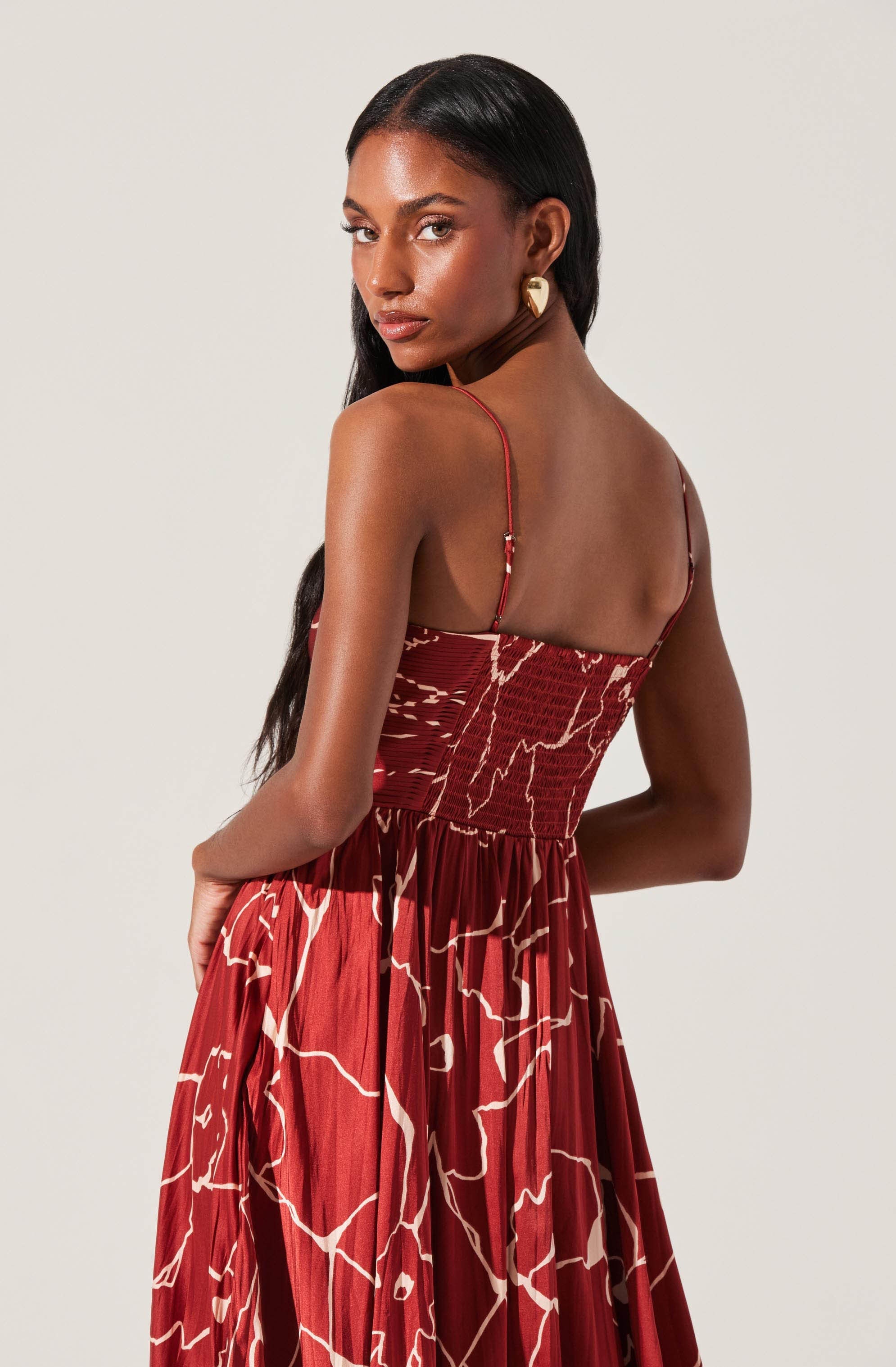 A woman with long, dark hair is wearing a sleeveless, red dress with thin spaghetti straps. The dress features a bold, abstract white and beige pattern resembling lightning or cracks, and has a fitted smocked bodice. The skirt is flowing and gathered at the waist. She is accessorized with gold geometric earrings.