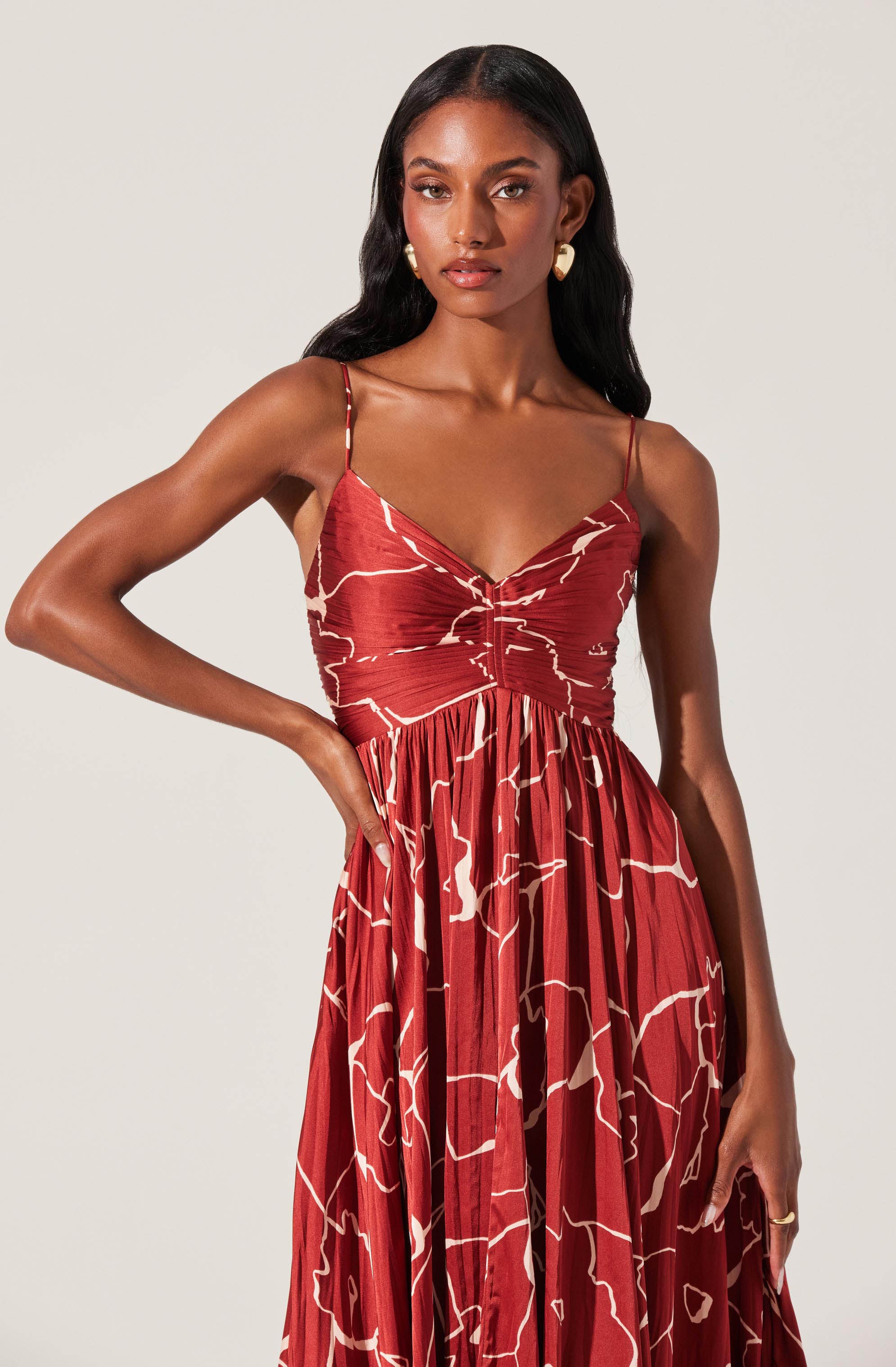 A woman is wearing a sleeveless, floor-length dress with thin spaghetti straps. The dress features a v-neckline and a fitted bodice, with a gathered and pleated skirt that flows down to the floor. The fabric has a red base color with an abstract, cream-colored, line-drawn pattern. She is accessorized with large, heart-shaped earrings and a ring on her right hand.