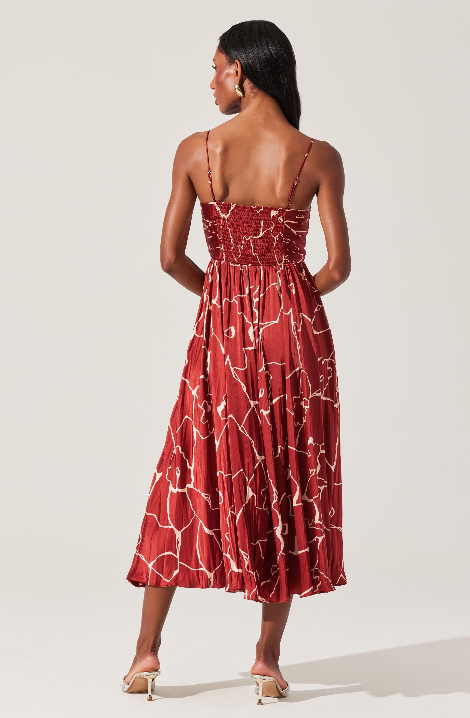 The dress features a fitted, smocked bodice with thin spaghetti straps and a flowy, pleated skirt that falls to mid-calf length. The fabric is red with an abstract, irregular white line pattern throughout.