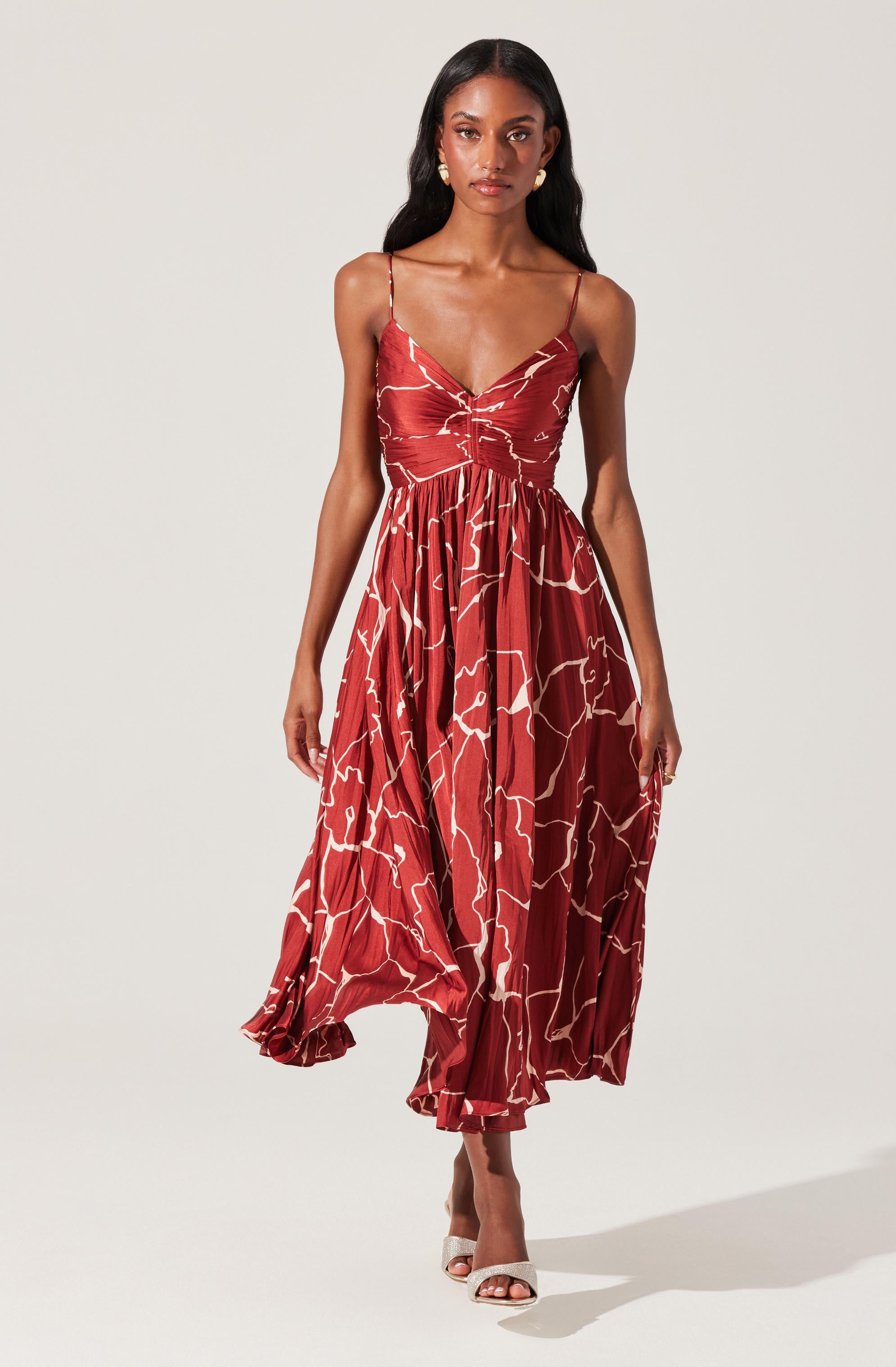 A woman is wearing a sleeveless, ankle-length dress with thin spaghetti straps. The dress has a deep V neckline and is fitted at the bodice with a ruched detail at the waist. The fabric features a red background with a white, irregular abstract pattern. She pairs the dress with white open-toe heels and gold earrings.