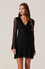 A woman is wearing a black dress made of lace with long, sheer sleeves featuring a striated pattern. The dress has a ruffled collar and a V-neckline with a bow detail at the front. The hemline of the dress is above the knees.